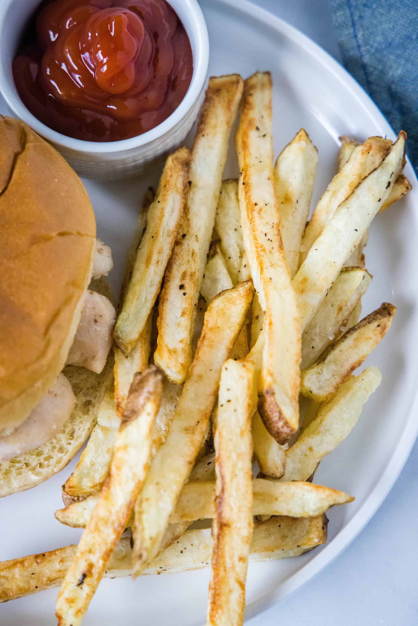 Homemade air fryer French fries are just as crispy as regular fries! Make them in a fraction of the time, with any seasonings you’d like. Overhead view of homemade air fryer French fries next to a burger and a small ramekin filled with ketchup on a plate.