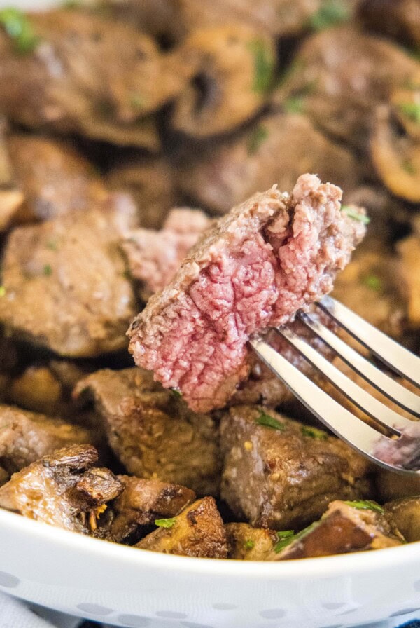 A piece of steak on a fork with a plate of steak and mushroom bites in the background