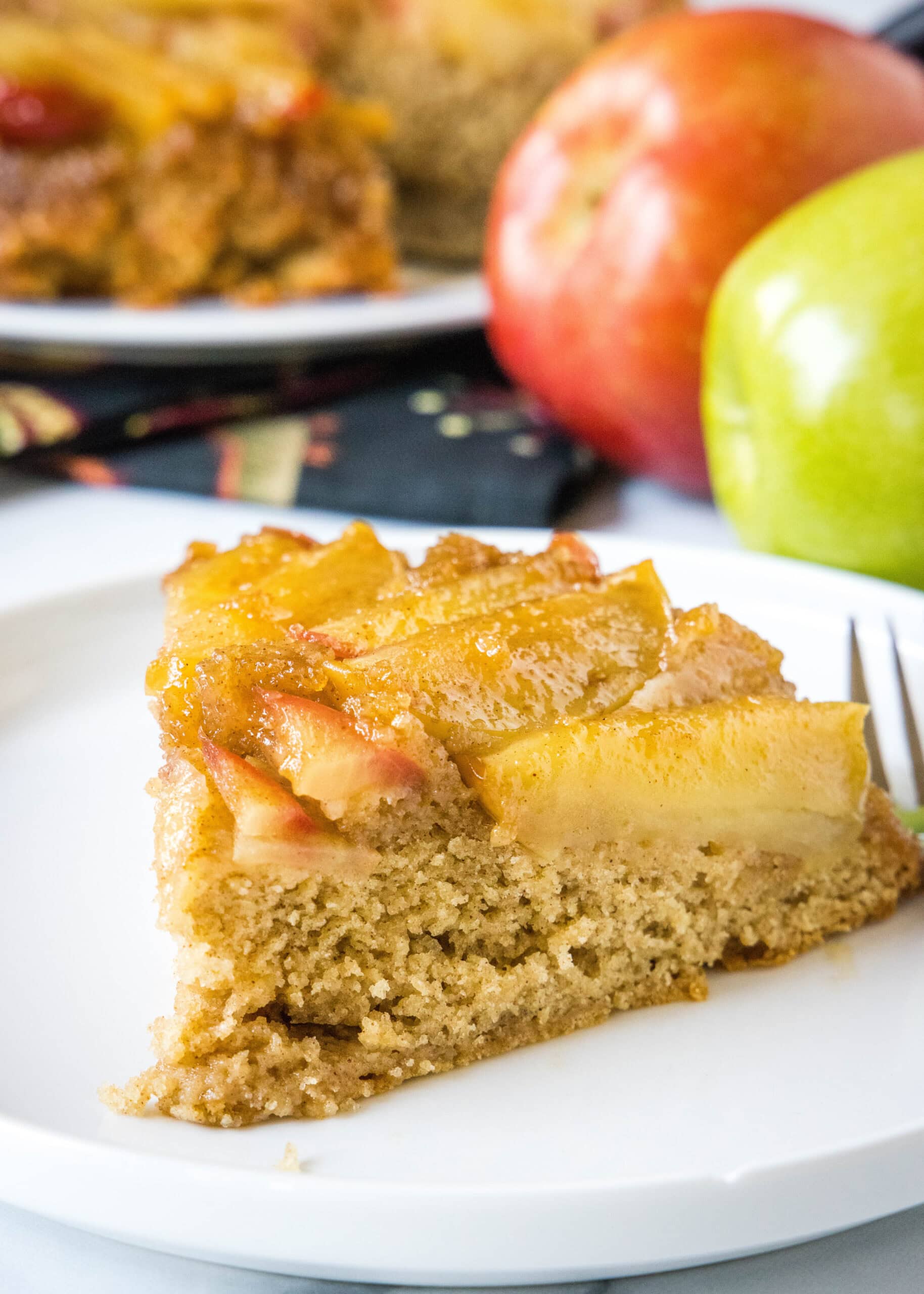 A slice of apple upside down cake on a white plate next to a fork, with red and green apples in the background.