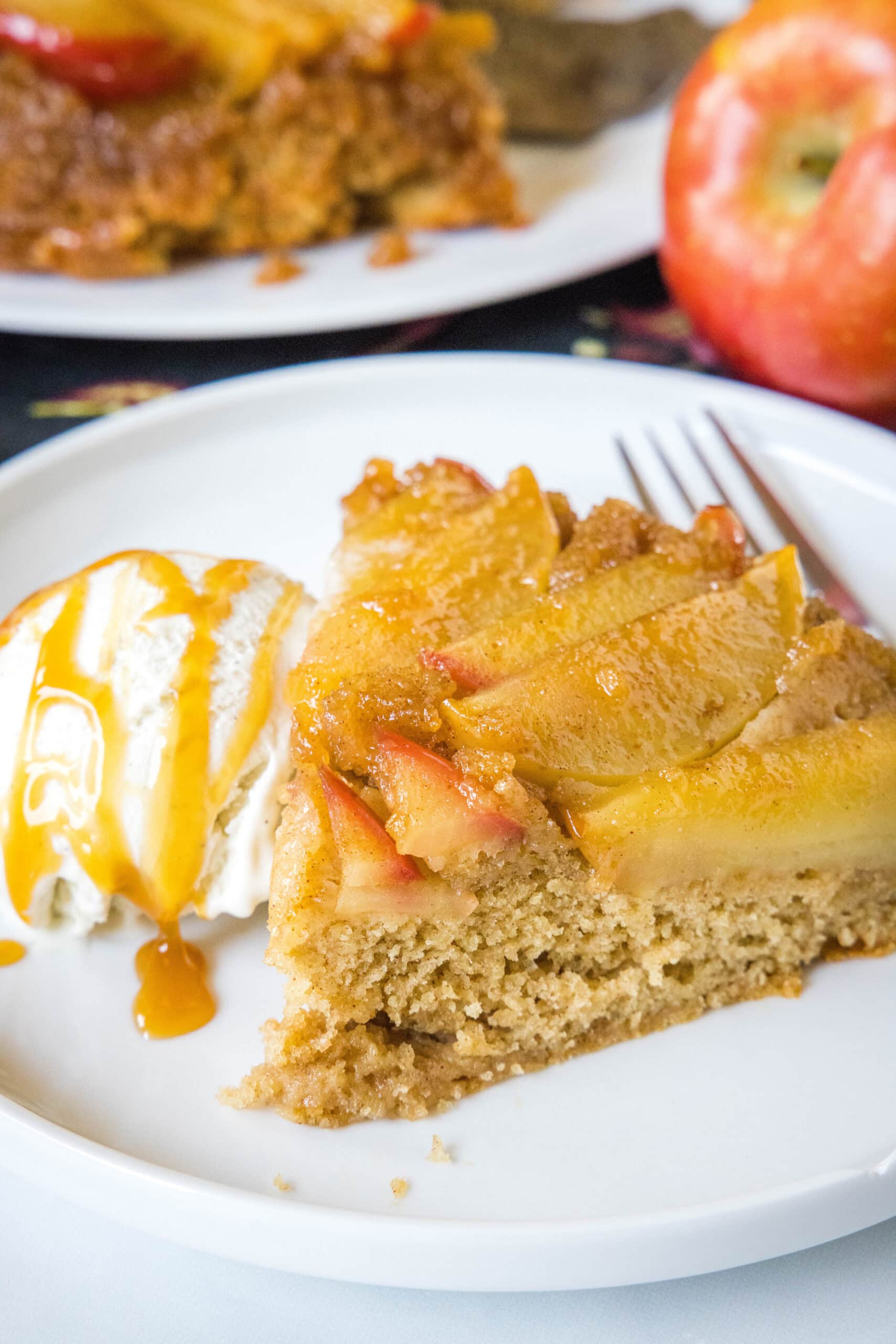 Apple upside down cake is a fall spin on a classic dessert, combining soft, spiced vanilla cake with a topping of gooey caramelized apples. A slice of apple upside down cake on a white plate next to a scoop of ice cream drizzled with caramel sauce, and a fork.
