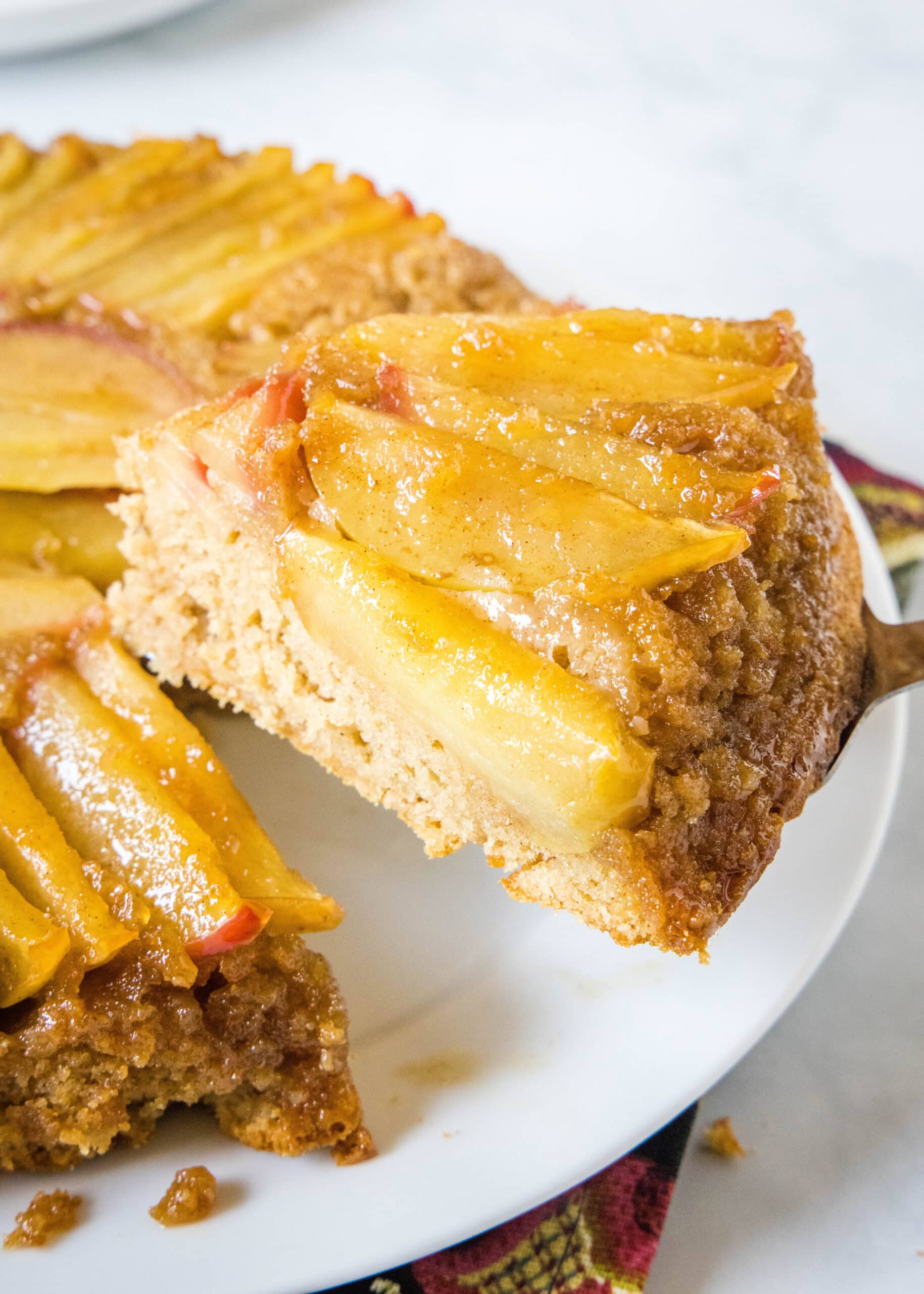 Close up of a slice of apple upside down cake being lifted from the rest of the cake on a white plate.
