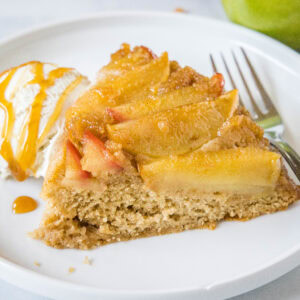 A slice of apple upside down cake on a white plate next to a scoop of ice cream drizzled with caramel sauce, and a fork.