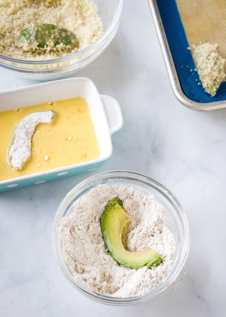Overhead view of a breading station for avocado fries, with avocado slices dipped in bowls of flour, egg, and breadcrumbs.