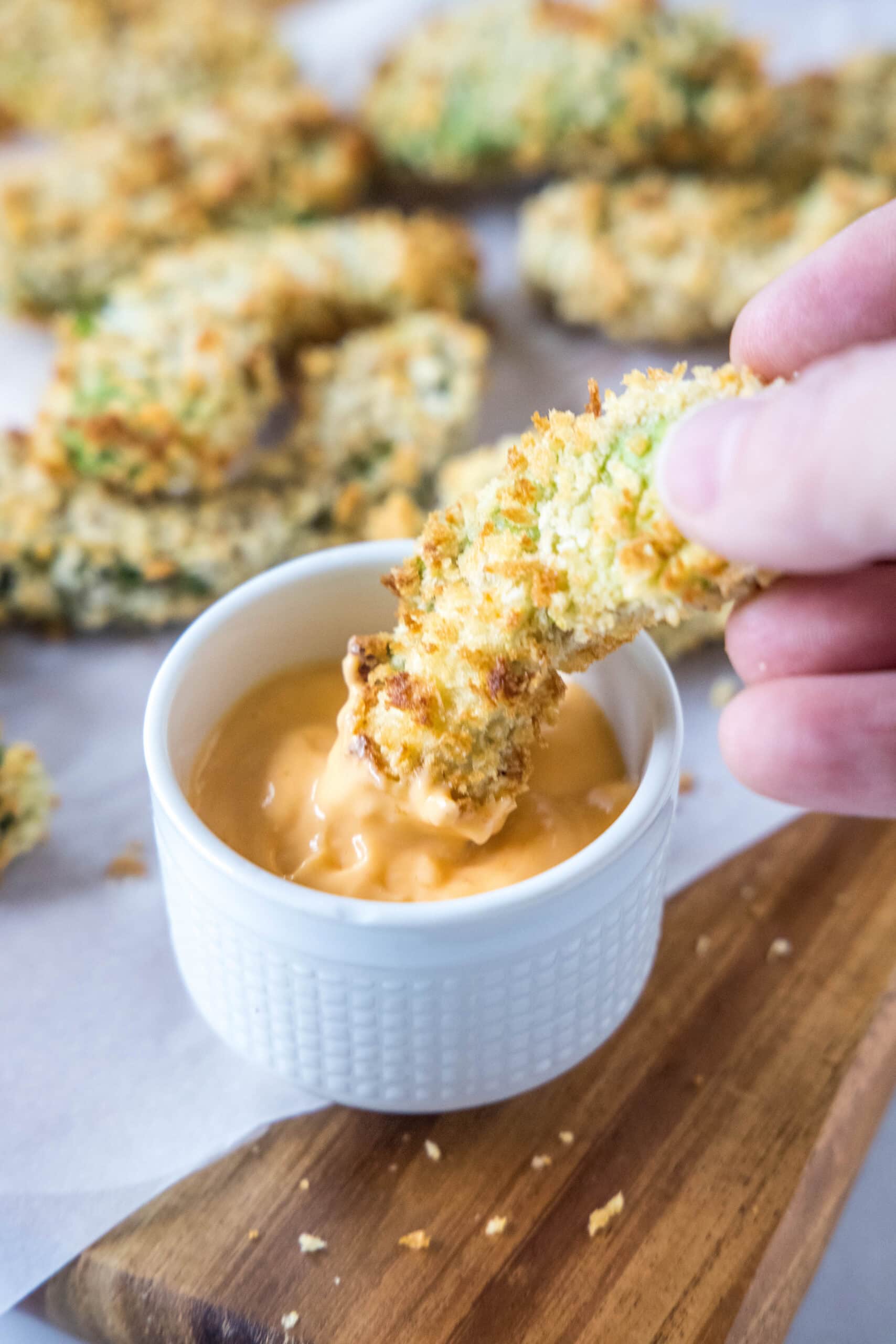 A hand dipping an avocado fry into a small bowl of dipping sauce, with more fries in the background.