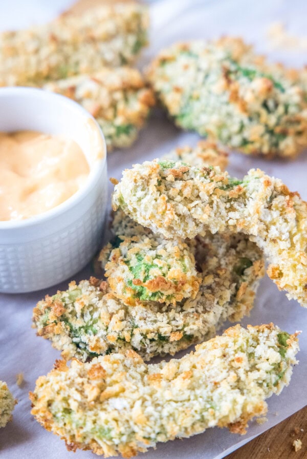 Avocado fries on a wooden cutting board lined with parchment paper, next to a small bowl of dipping sauce.