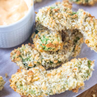 Avocado fries on a wooden cutting board lined with parchment paper, next to a small bowl of dipping sauce.