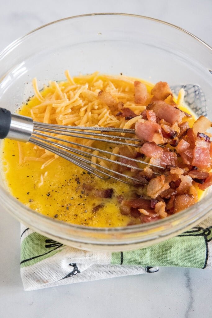 Crumbled bacon and cheese added to a bowl of beaten eggs with a whisk.