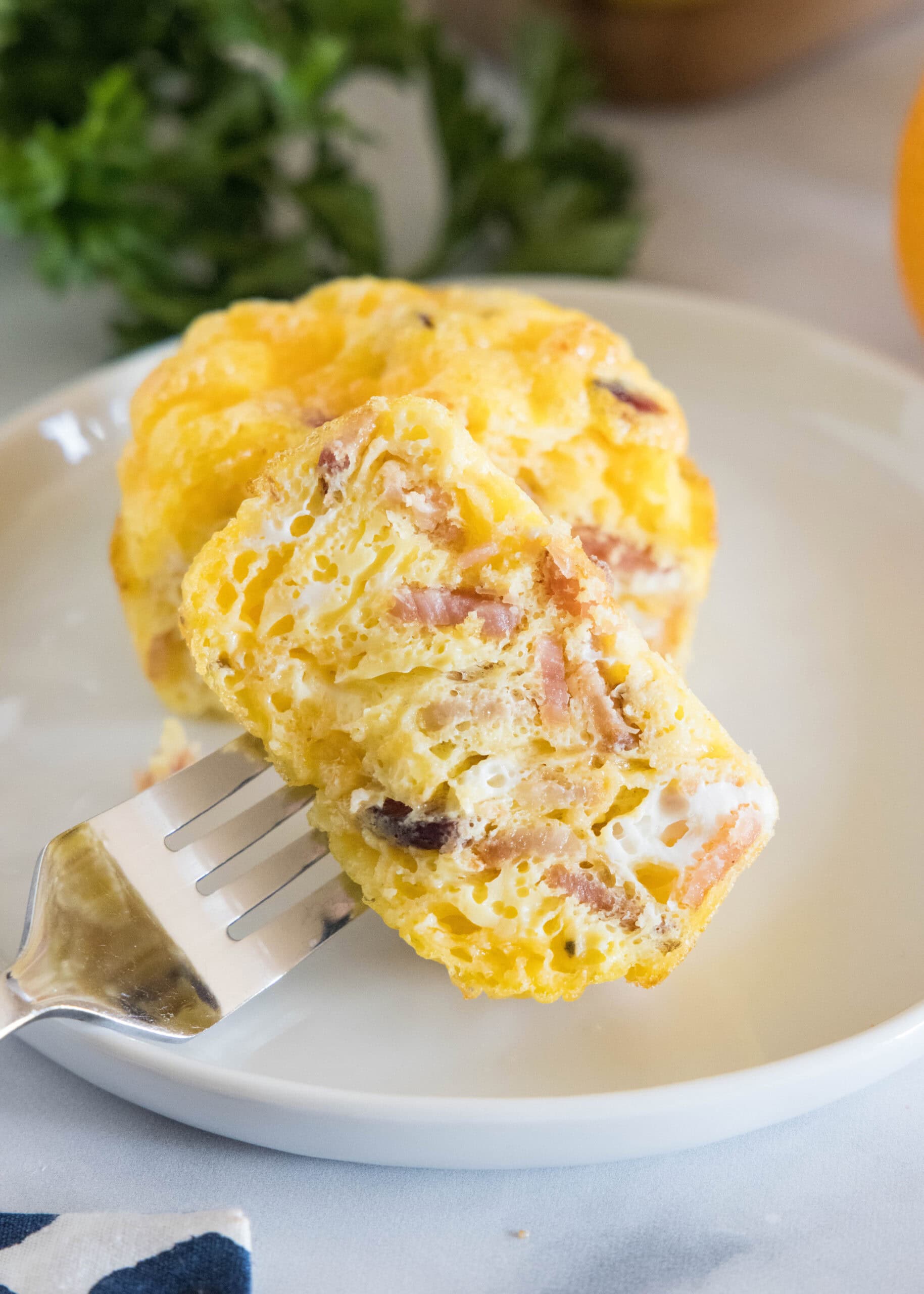 A fork stuck into one half of a bacon cheese egg muffin on a white plate.