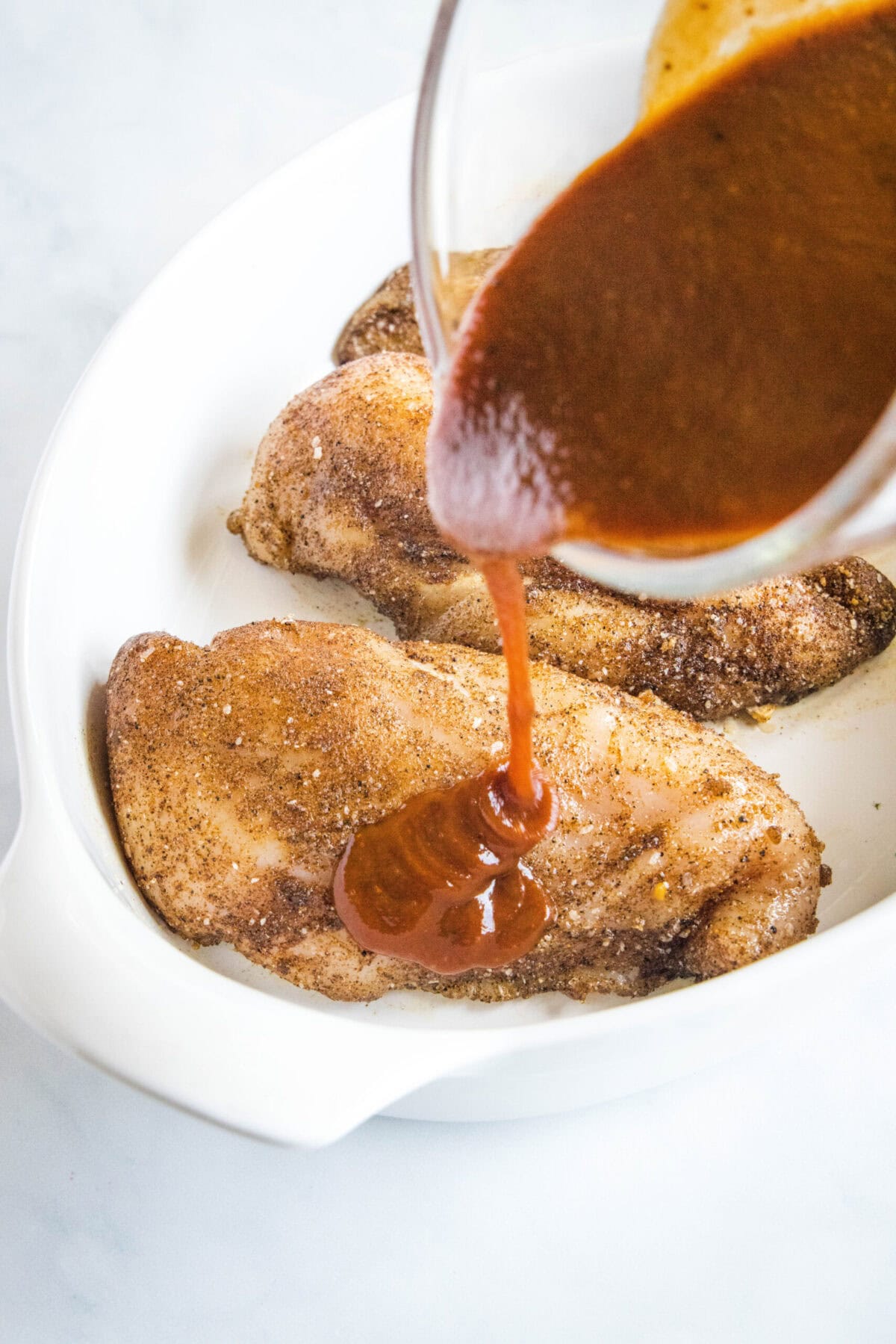 Barbecue sauce being poured over three seasoned, raw chicken breasts in a dish