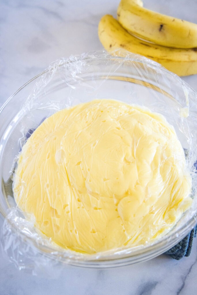 A mixing bowl with banana cream pie filling in it, covered in plastic wrap