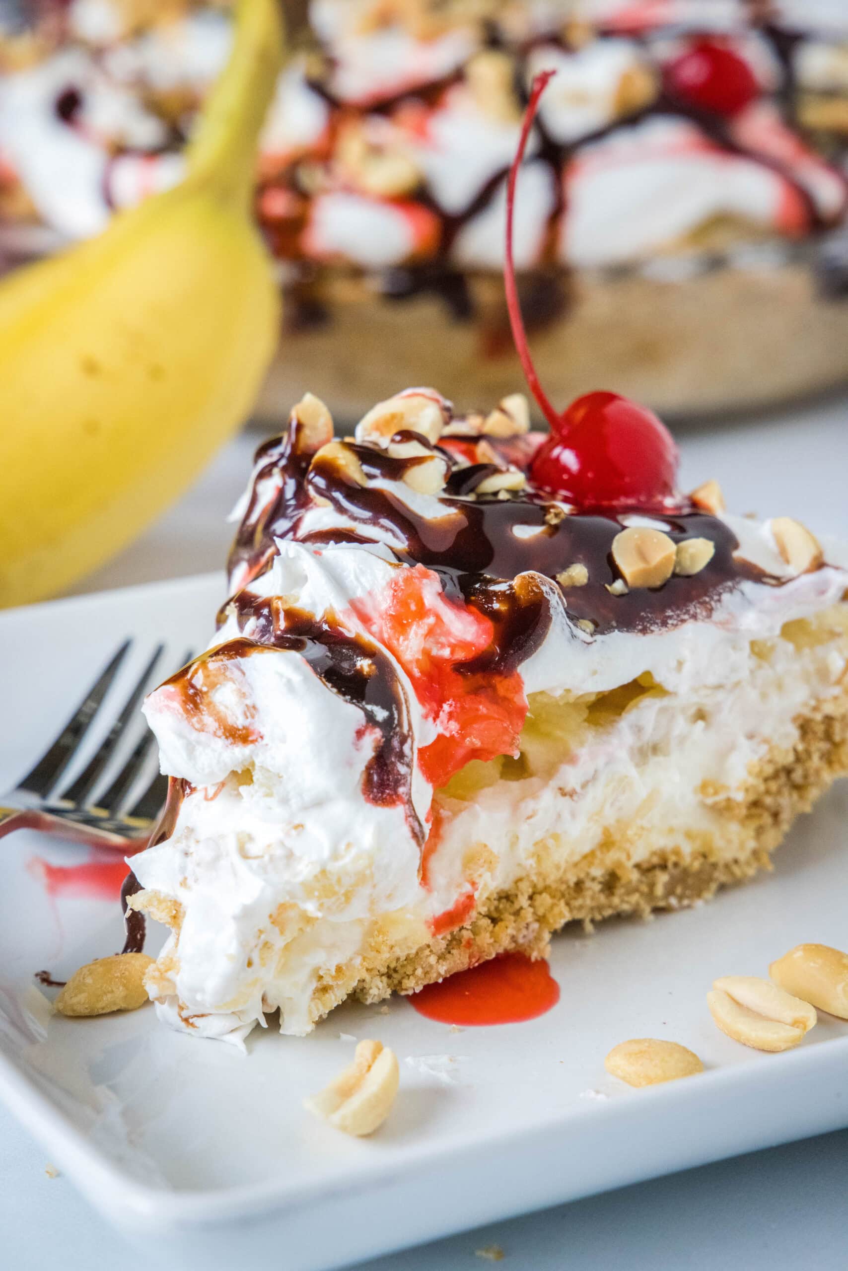 A slice of banana split pie drizzled with chocolate sauce and strawberry sauce, topped with chopped peanuts and cherries on a plate, with a banana and the rest of the pie in the background.