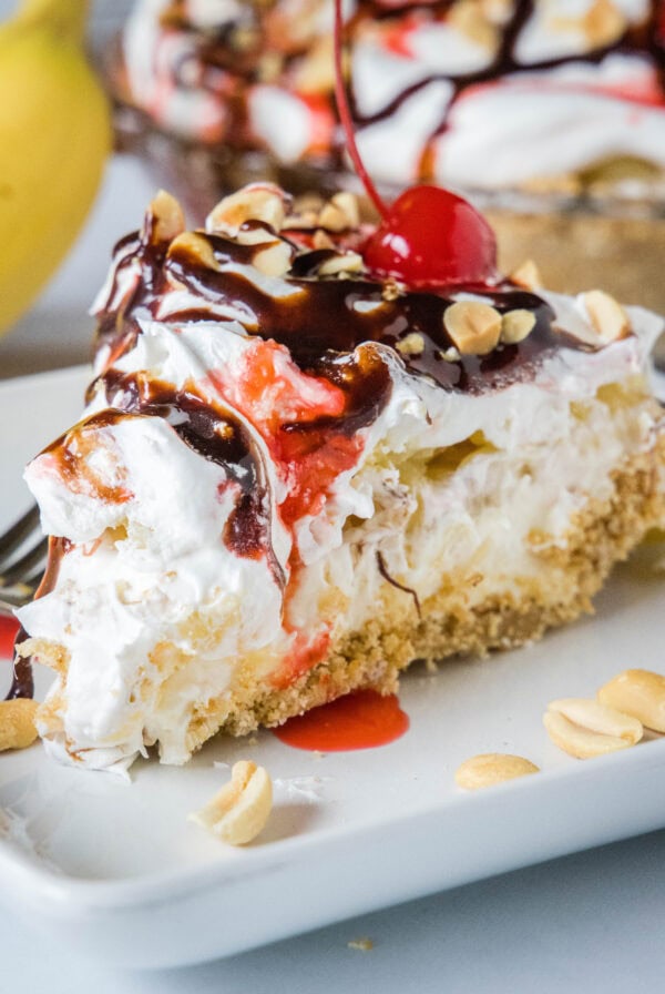 A slice of banana split pie drizzled with chocolate sauce and strawberry sauce, topped with chopped peanuts and cherries on a plate.