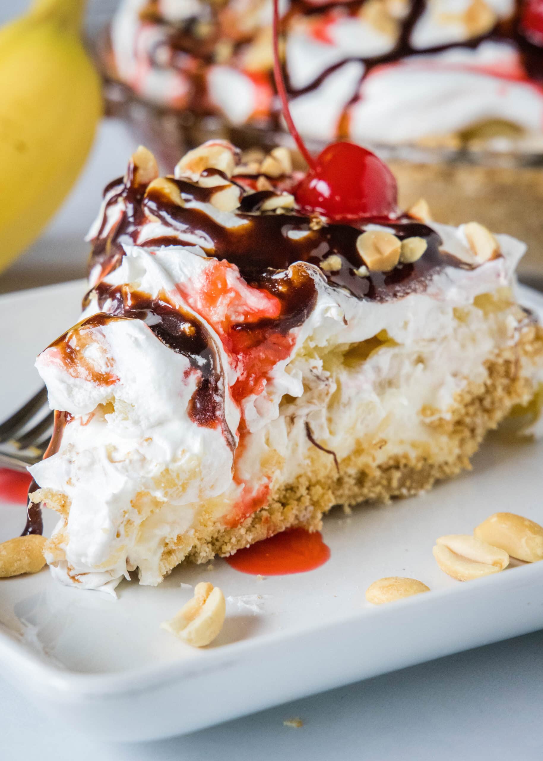 Banana split pie is a cool and creamy no-bake dessert that you can make in 20 minutes. Filled with whipped cream, bananas, and pineapple! A slice of banana split pie drizzled with chocolate sauce and strawberry sauce, topped with chopped peanuts and cherries on a plate.
