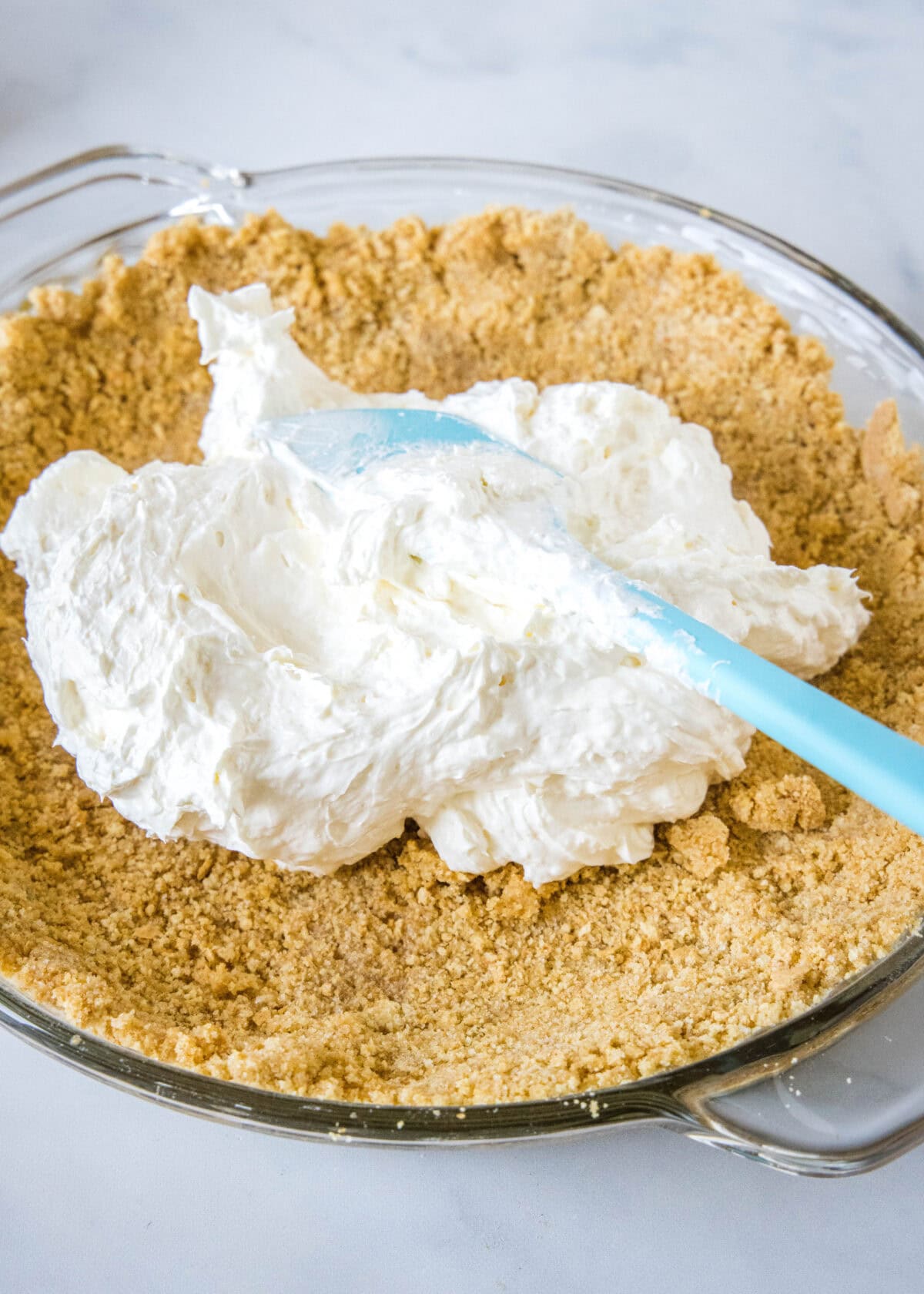 Cream cheese pie filling added to a graham cracker crust with a blue spatula.