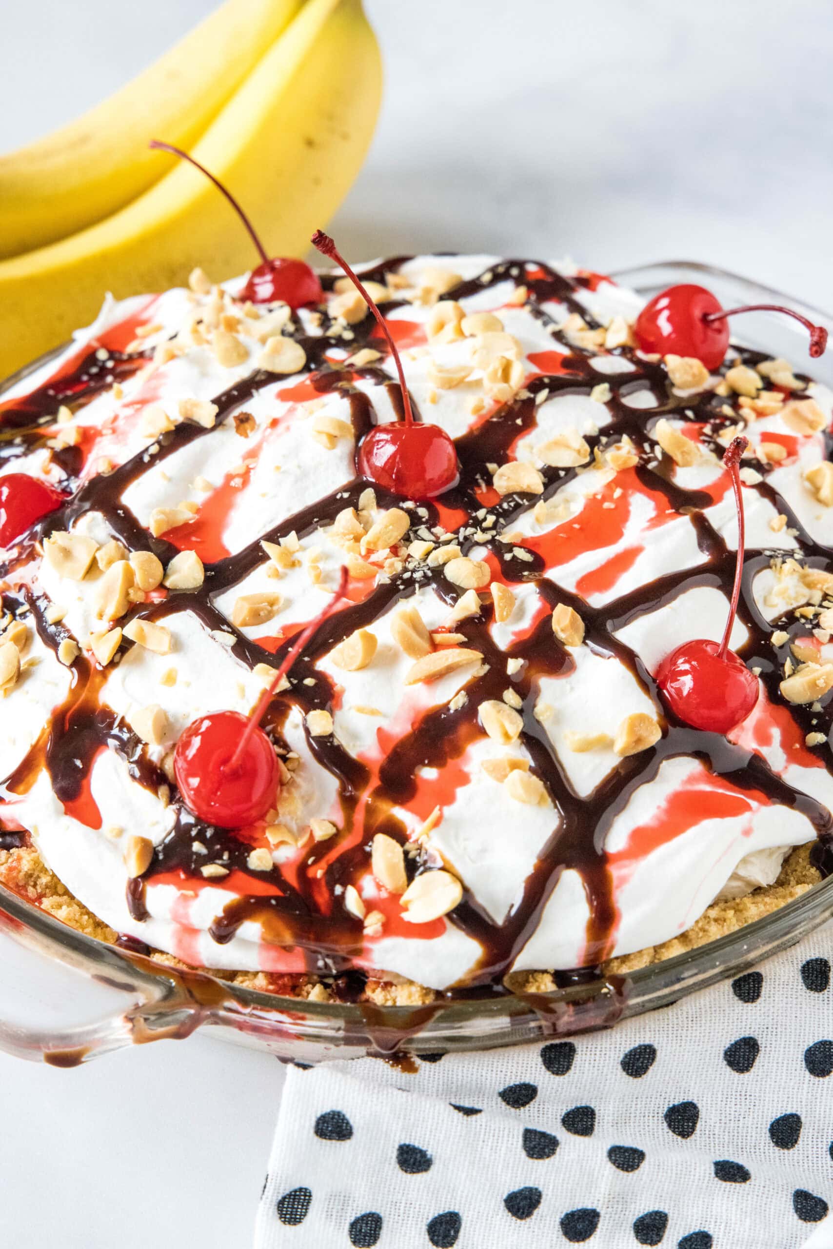 Banana split pie drizzled with chocolate sauce and strawberry sauce, topped with chopped peanuts and cherries.