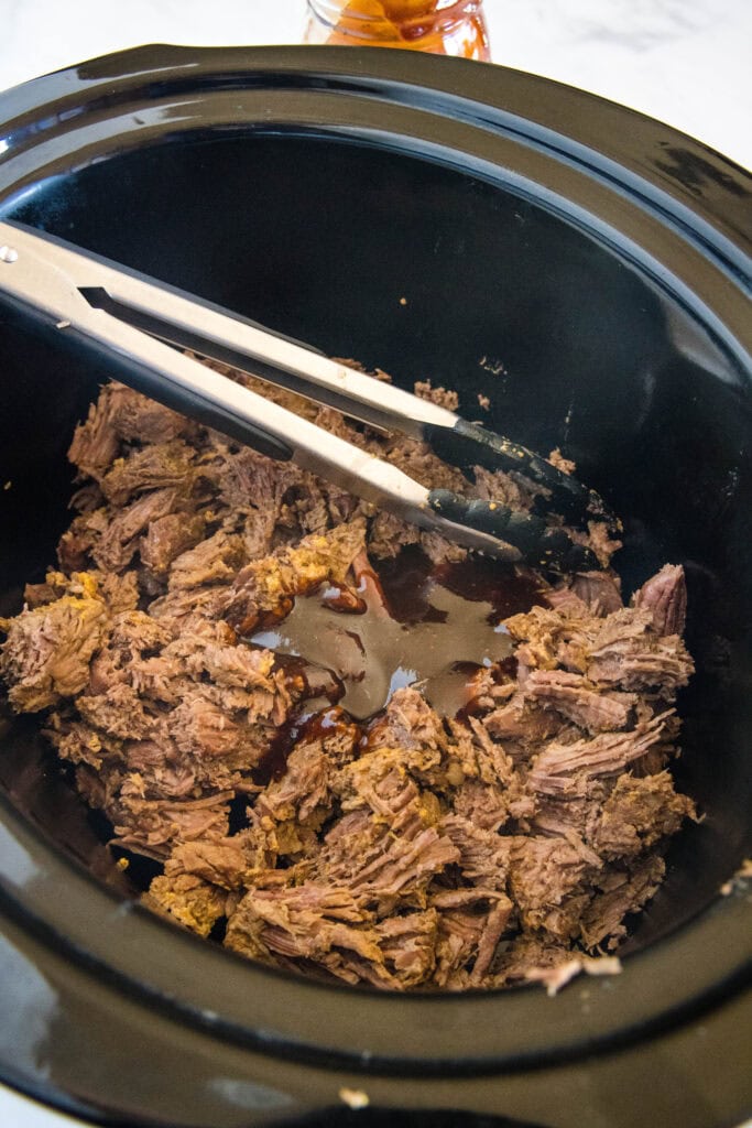 Metal tongs resting in shredded BBQ beef inside the crock pot.