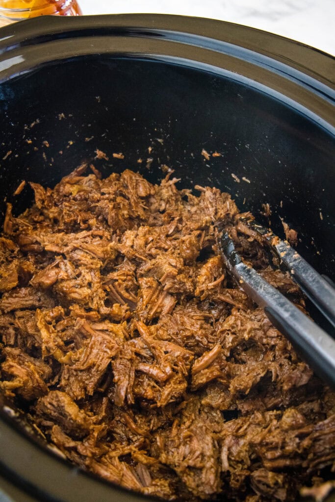 Metal tongs resting in shredded BBQ beef inside the crock pot.