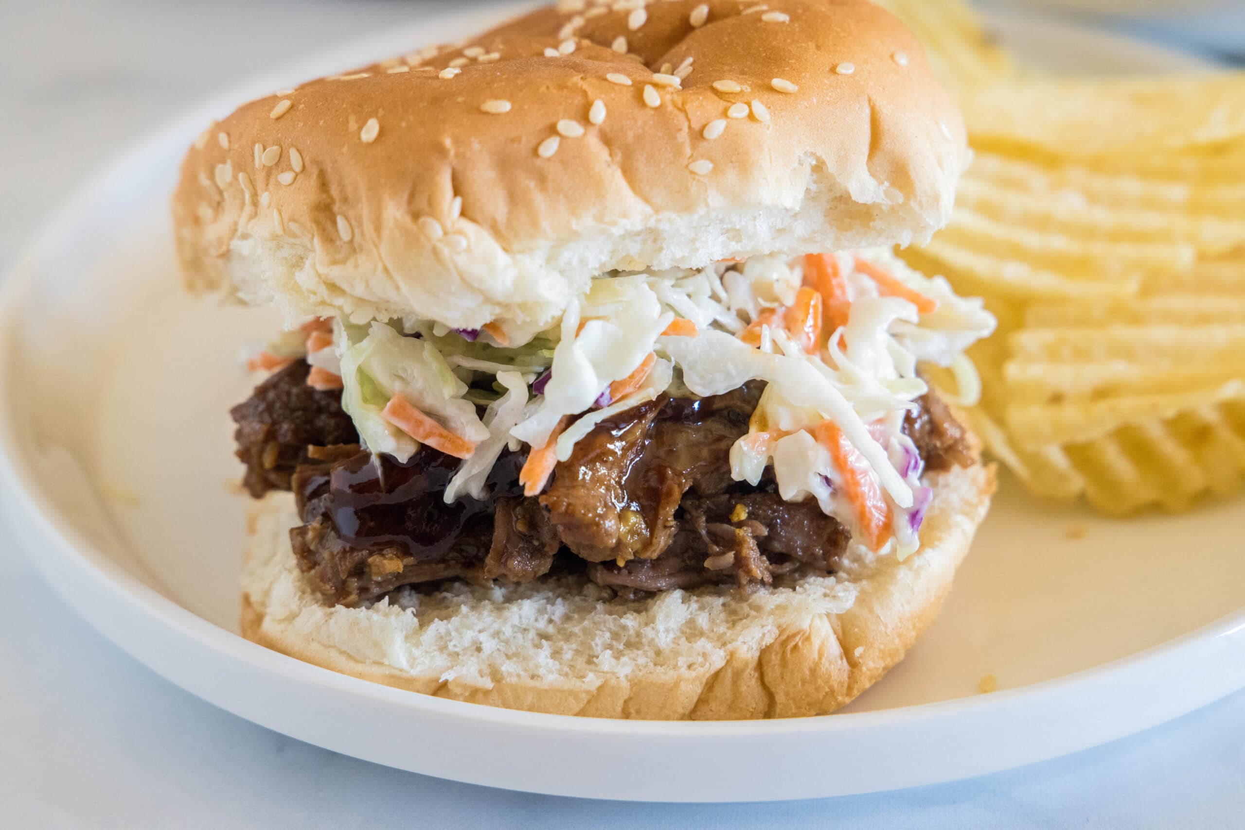 A BBQ beef sandwich with coleslaw, next to potato chips on a white plate.