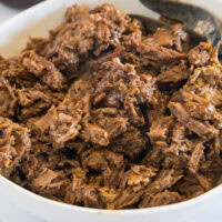 Metal tongs resting in shredded BBQ beef inside the crock pot.