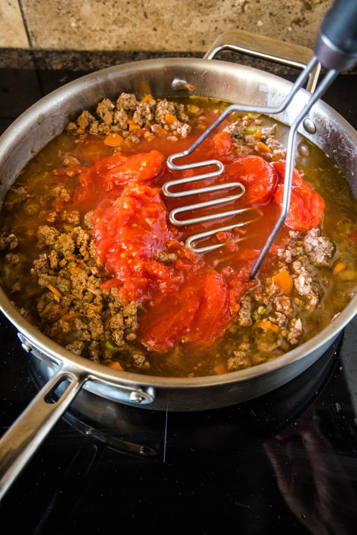 Tomatoes being crushed into meat sauce with a potato masher.