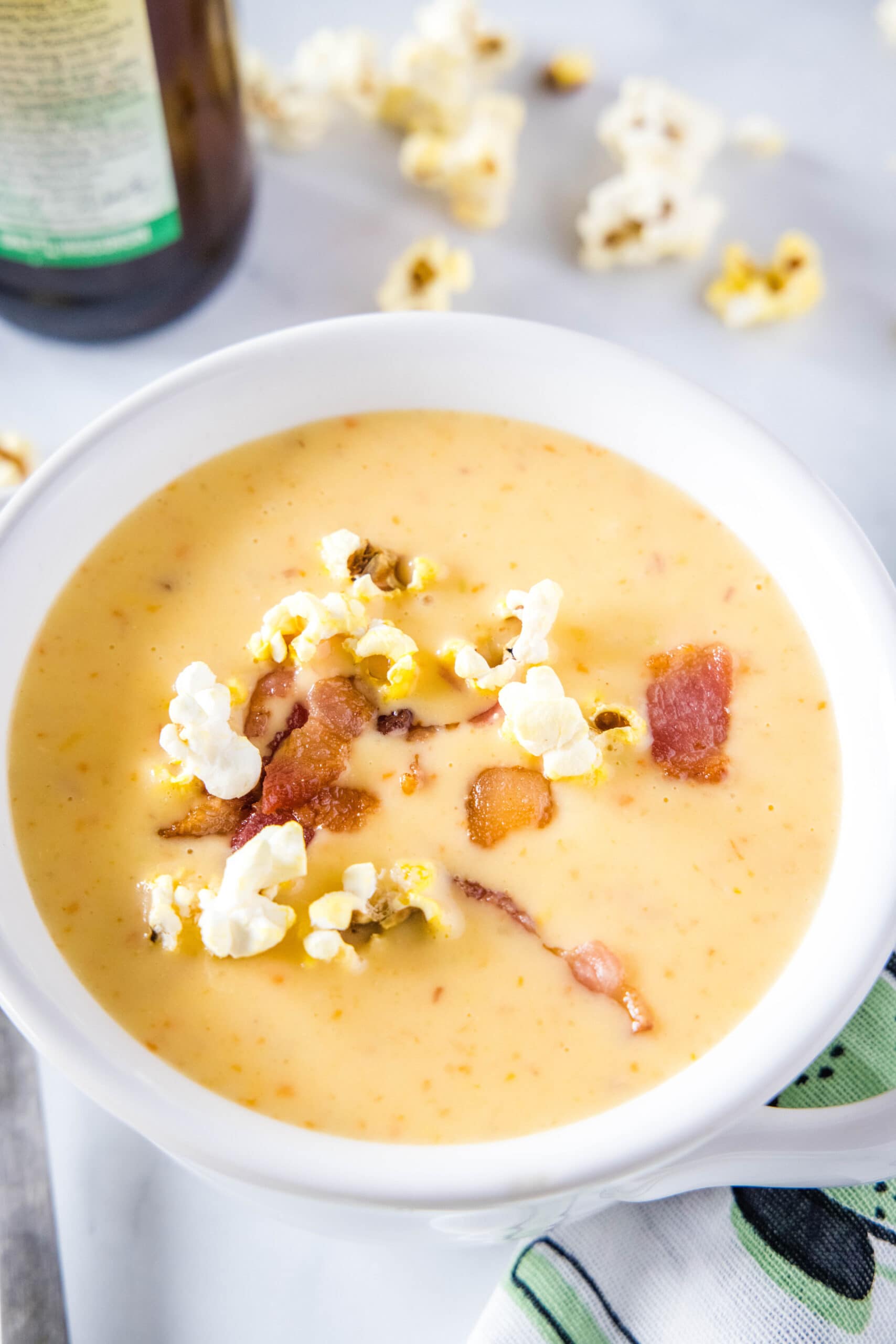 This hearty beer cheese soup is rich, creamy, and packed full of beer, cheddar cheese, and bacon. It's perfect for a cold night. A bowl of cheese soup topped with popcorn and bacon, with a bottle of beer and some popcorn in the background