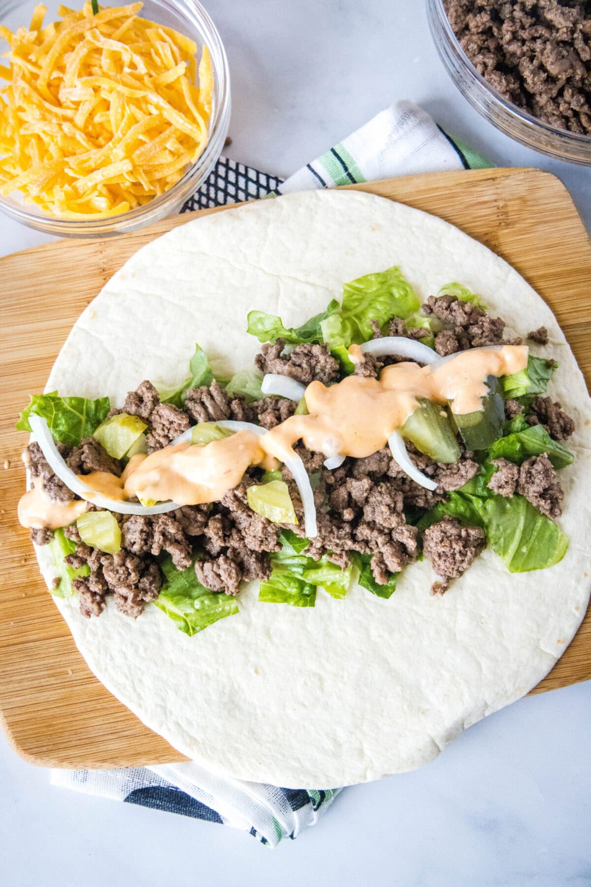 Overhead view of a flat tortilla topped with veggies, ground beef, and sauce, next to a bowl of cheese
