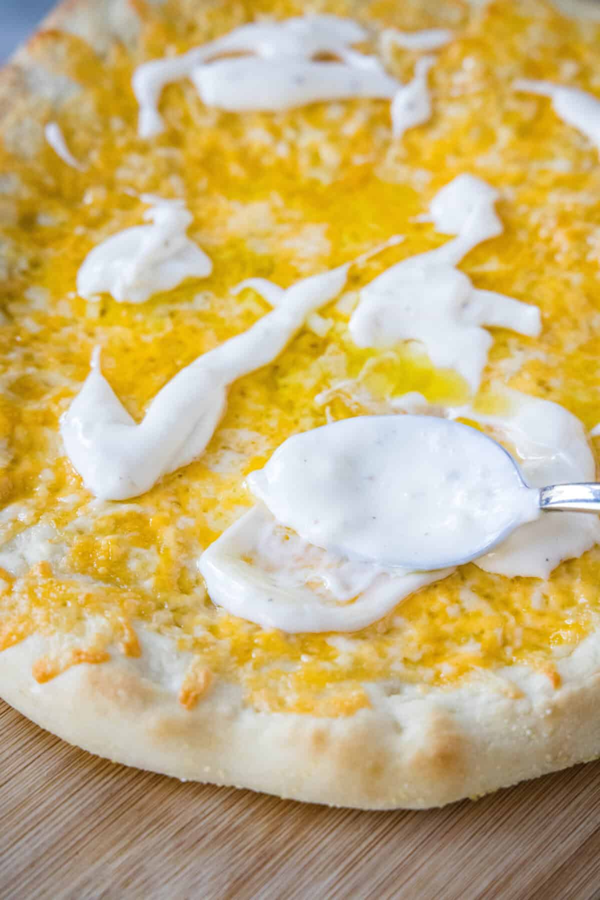 A spoon spreading aioli over melted cheese on top of a baked pizza crust.