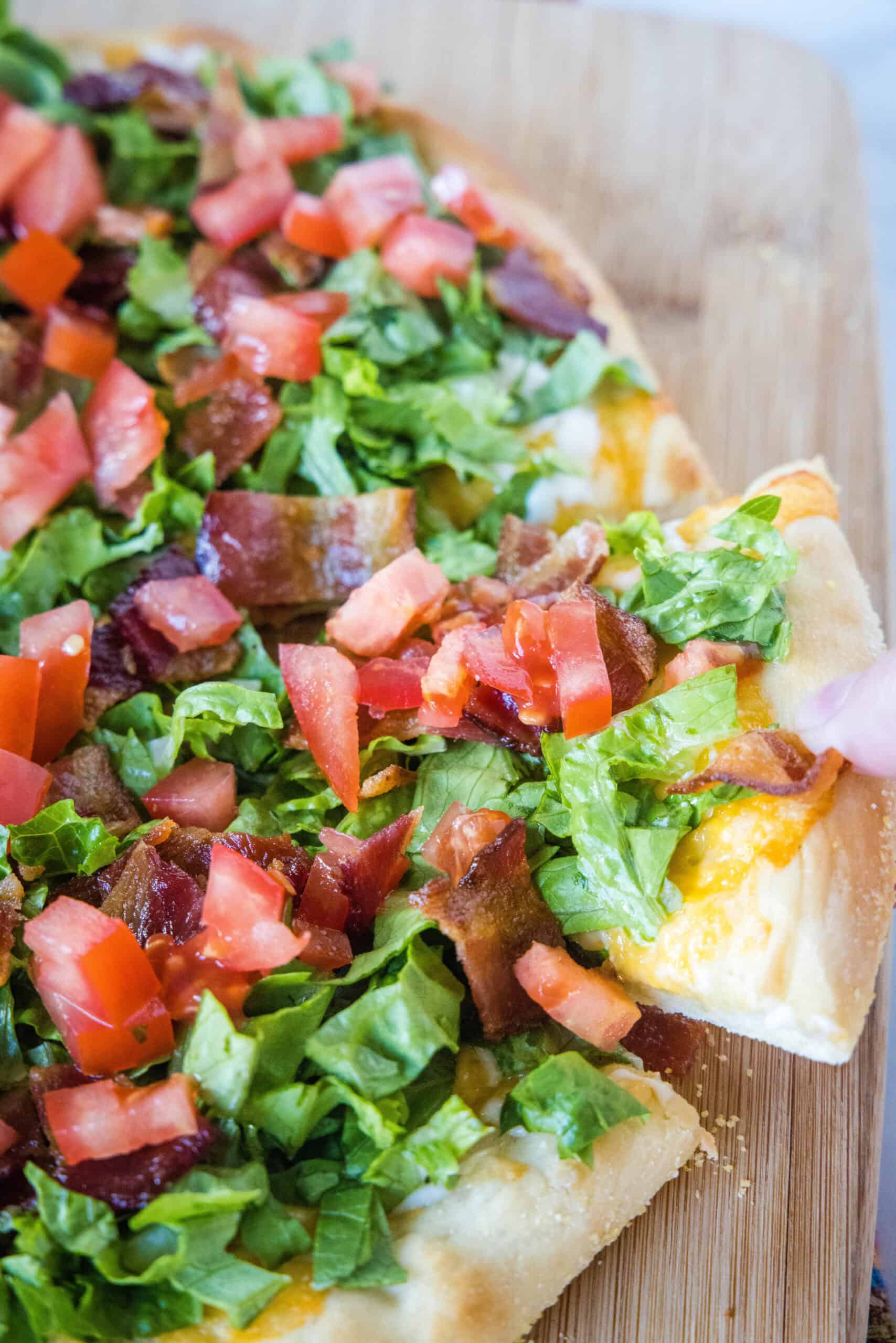 A hand lifting a slice from a BLT pizza on a wooden cutting board.