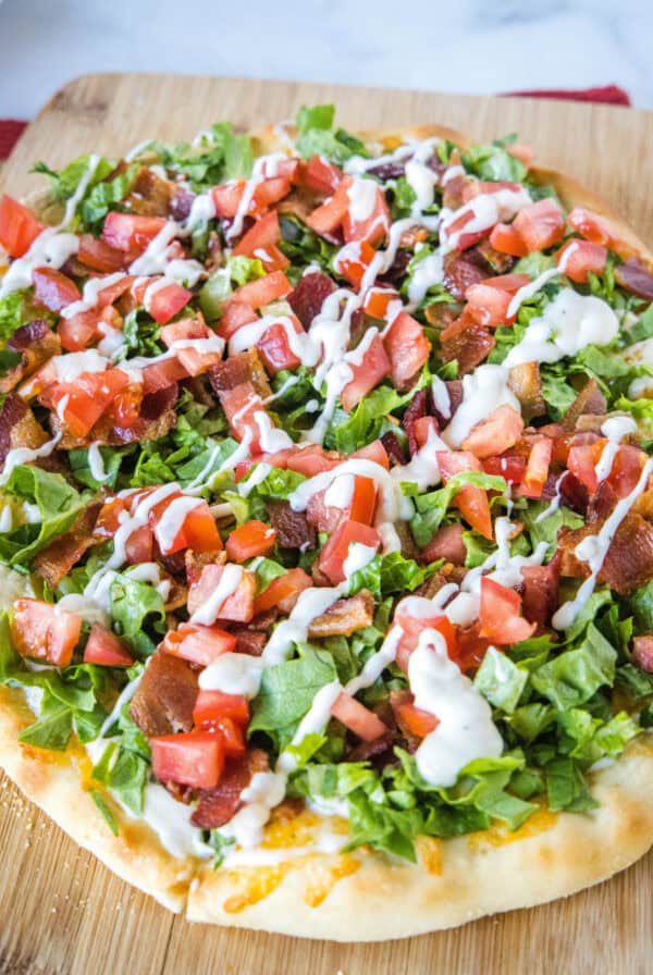 Close up of a BLT pizza topped with a drizzle of aioli on a wooden cutting board.