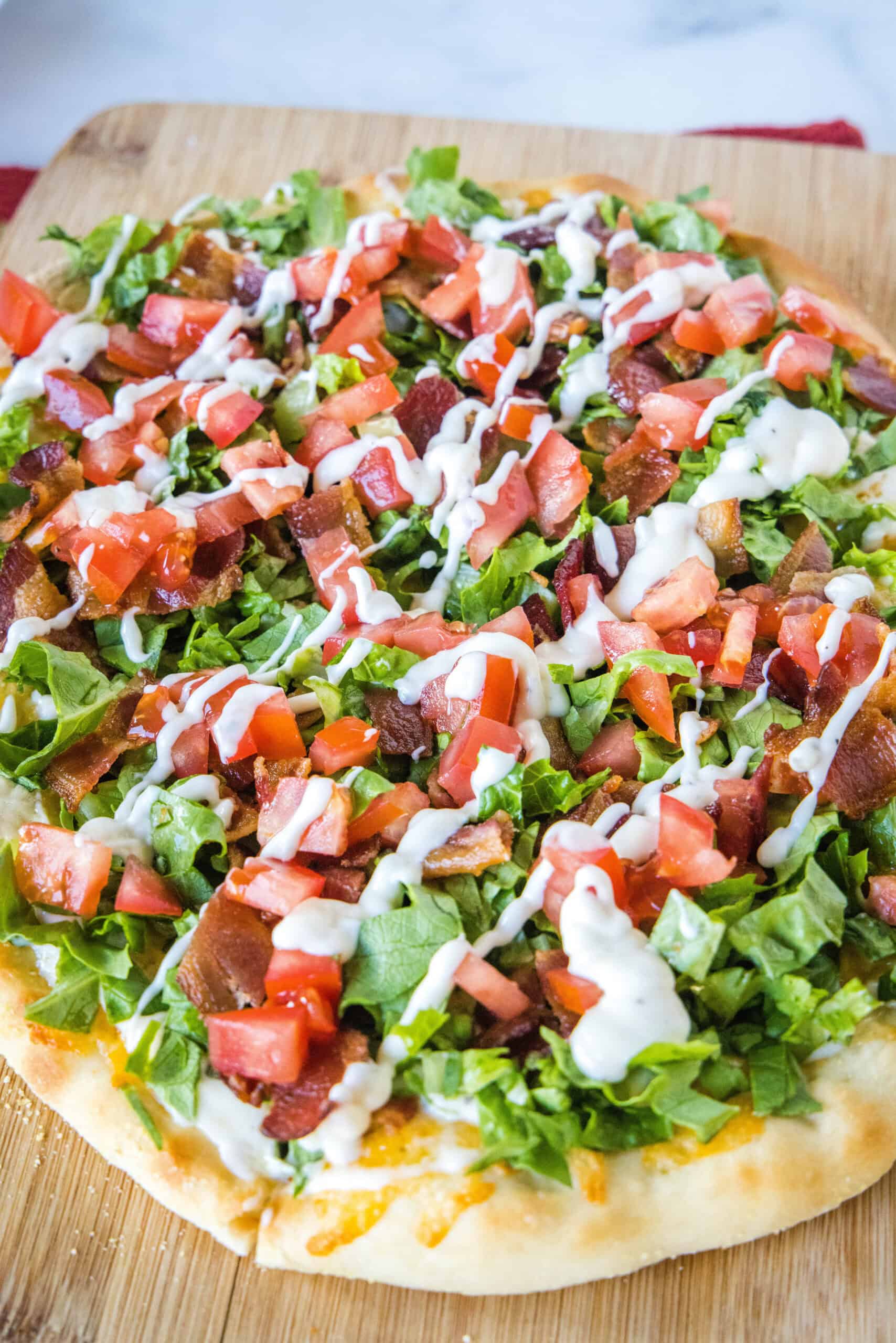 Close up of a BLT pizza topped with a drizzle of aioli on a wooden cutting board.