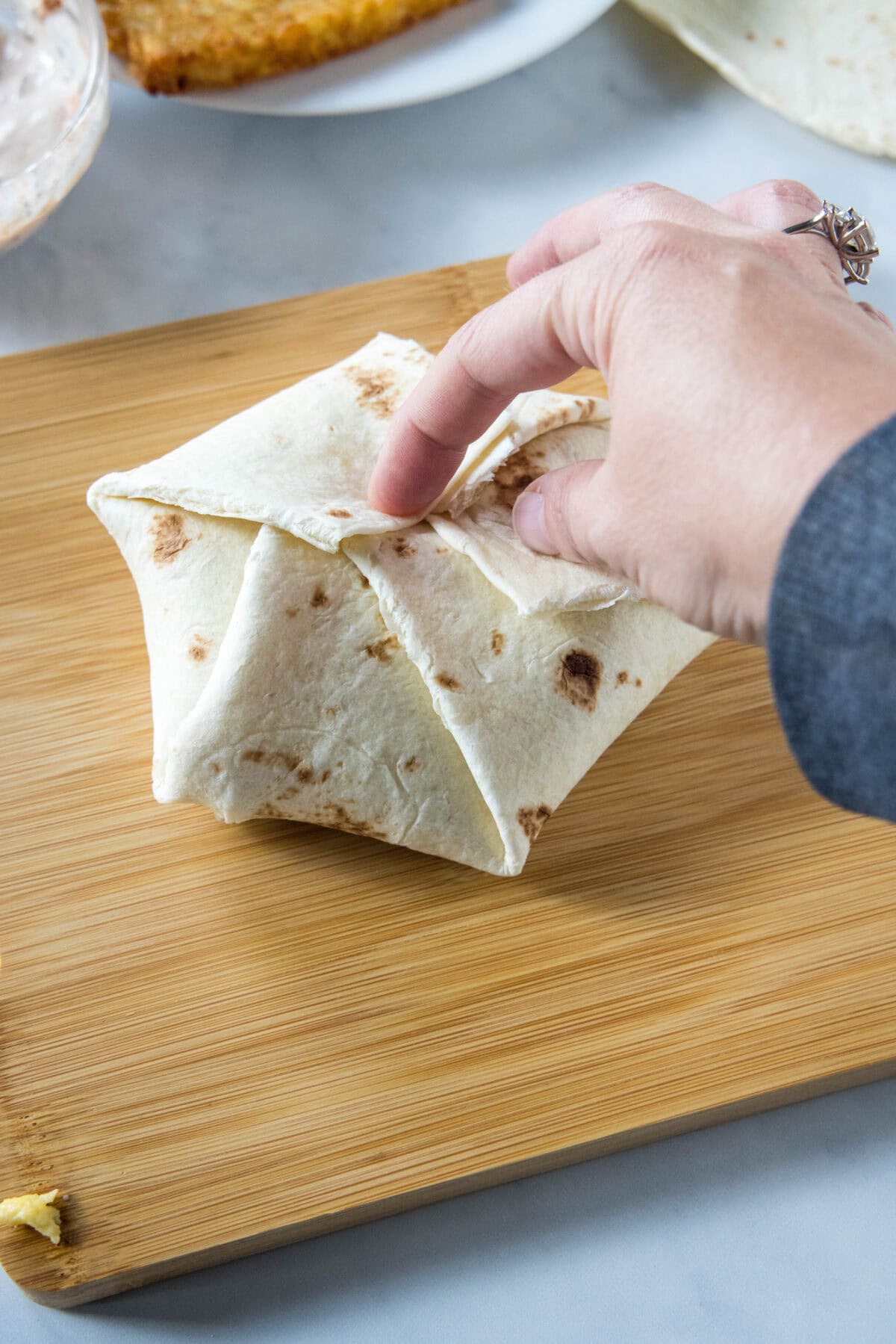 A hand folding a flour tortilla into a crunchwrap on a wooden cutting board.