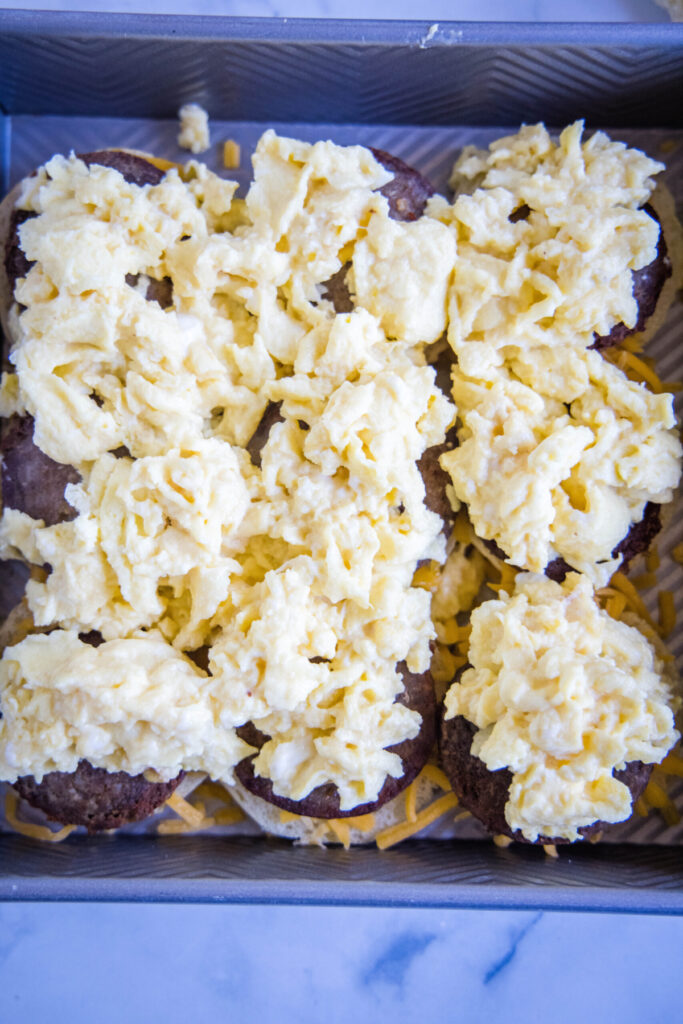 Overhead view of a baking tray with scrambled eggs on top of sausage on top of slider buns