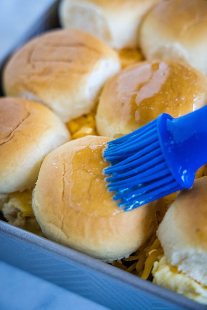 Butter being brushed onto the top of slider buns in a baking tray