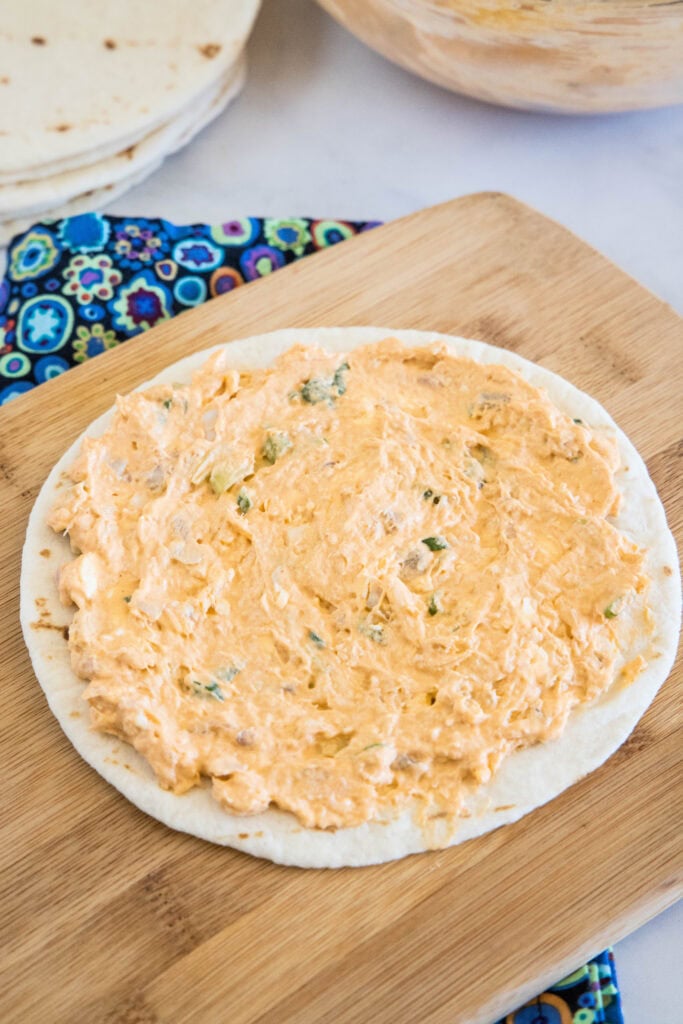 Buffalo chicken filling spread over a soft tortilla on a wooden cutting board.