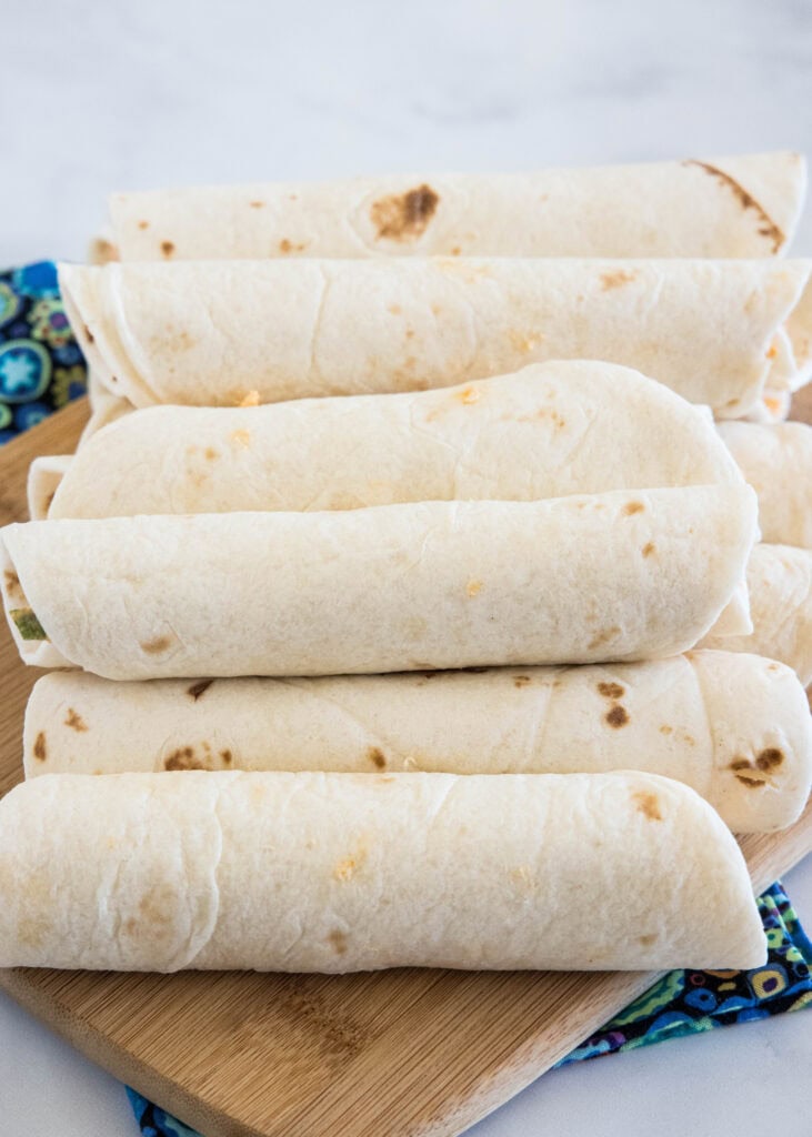 Rolled Buffalo chicken tortillas stacked on a wooden cutting board.