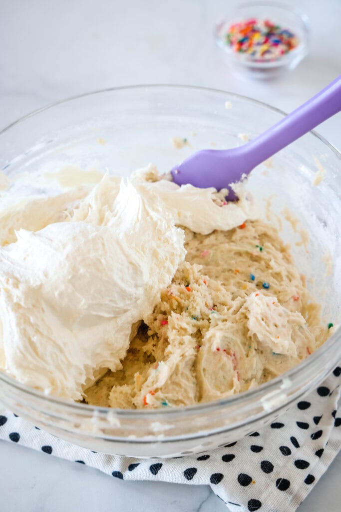 Cool whip partially folded into cake batter dip ingredients in a glass bowl with a purple rubber spatula.