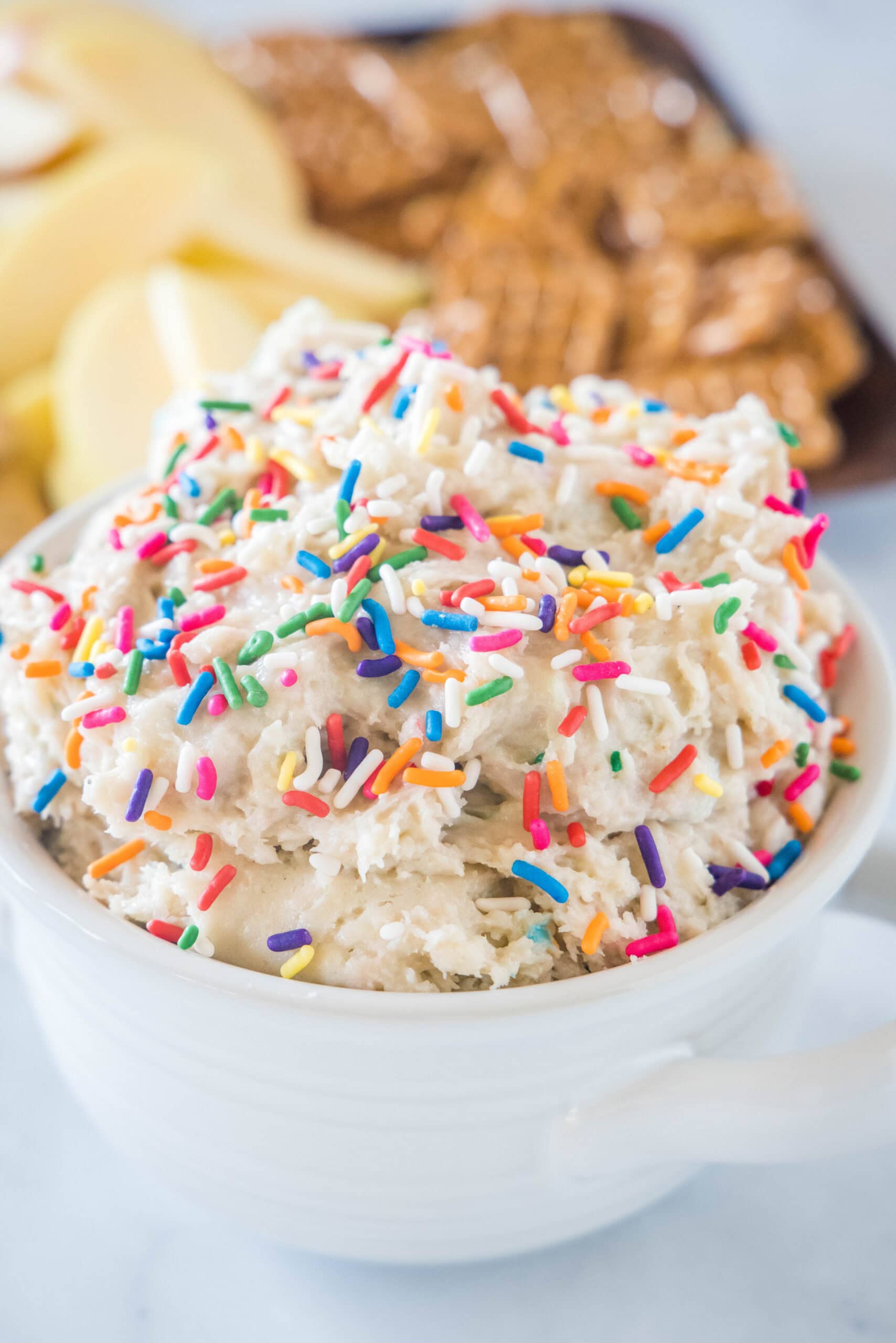 Cake batter dip in a white bowl topped with sprinkles, with a platter of cookies in the background.
