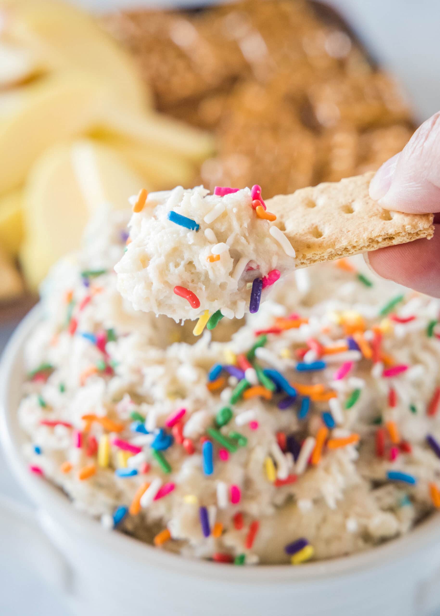 A hand holding a cookie over a bowl of cake batter dip, with a platter of cookies in the background.