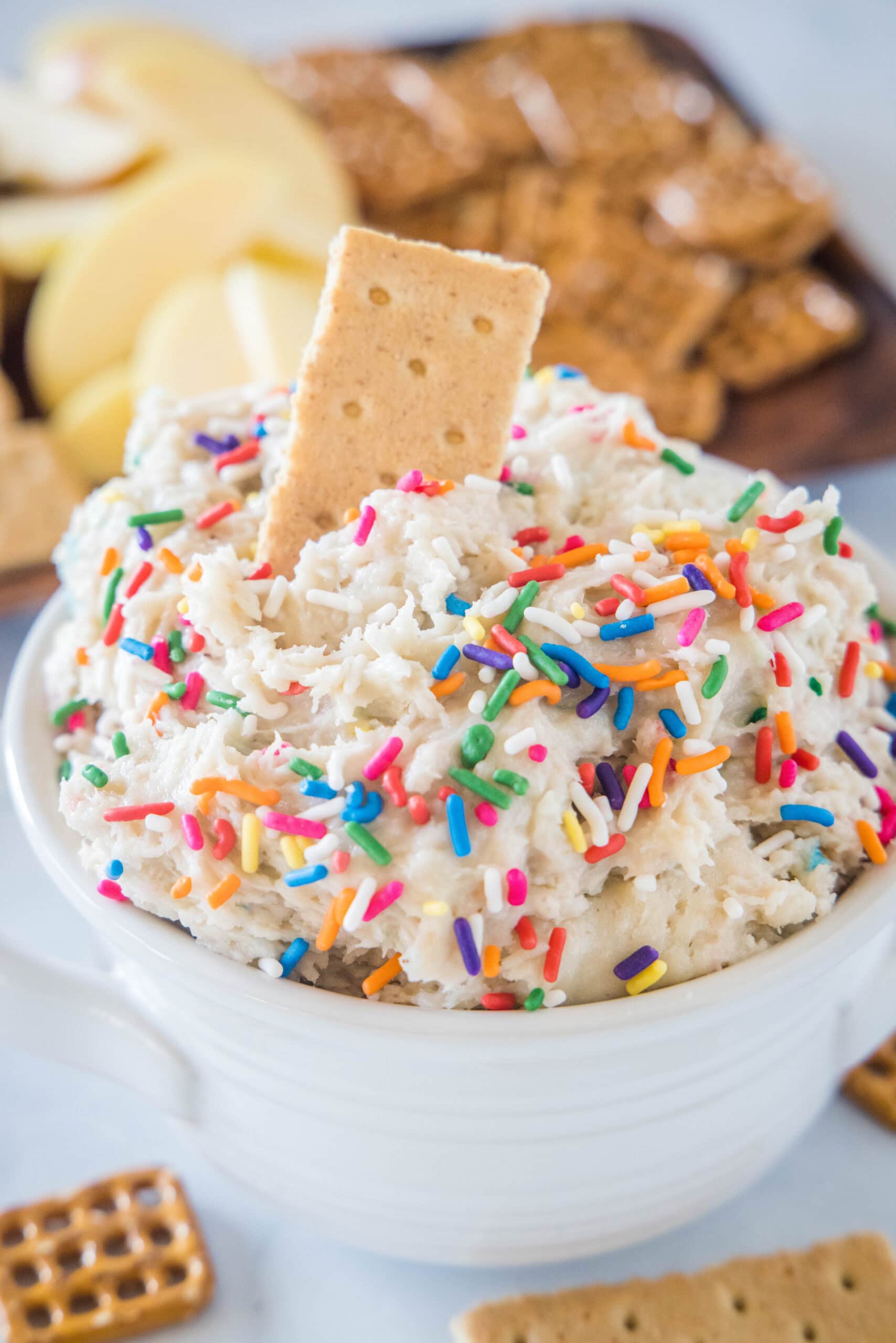 Cake batter dip in a white bowl with a cookie stuck into the top, and a platter of cookies in the background.