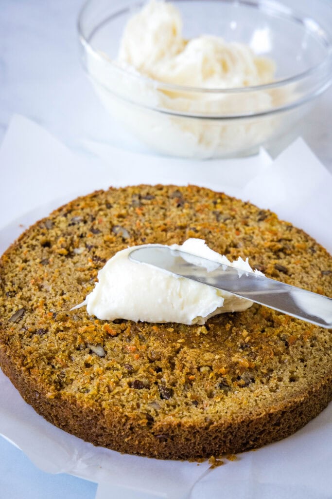 An offset spatula spreading cream cheese frosting over a carrot cake layer, with a glass bowl of frosting in the background.