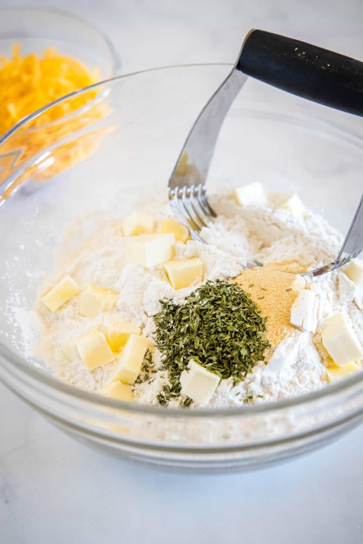 Cubed butter and herbs added to a bowl of flour, next to a pastry cutter.