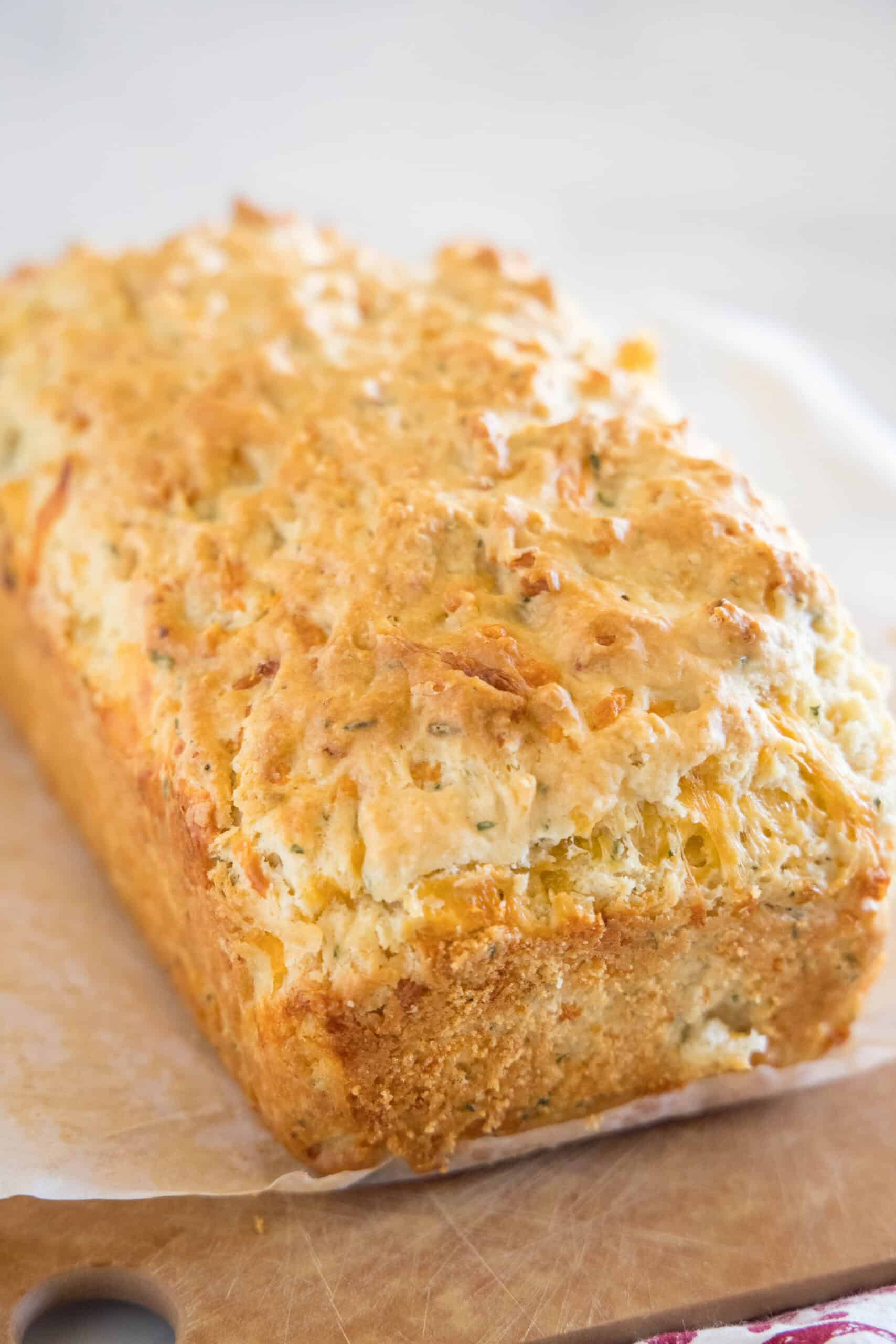 A loaf of cheesy quick bread on a piece of parchment paper on top of a wooden cutting board.