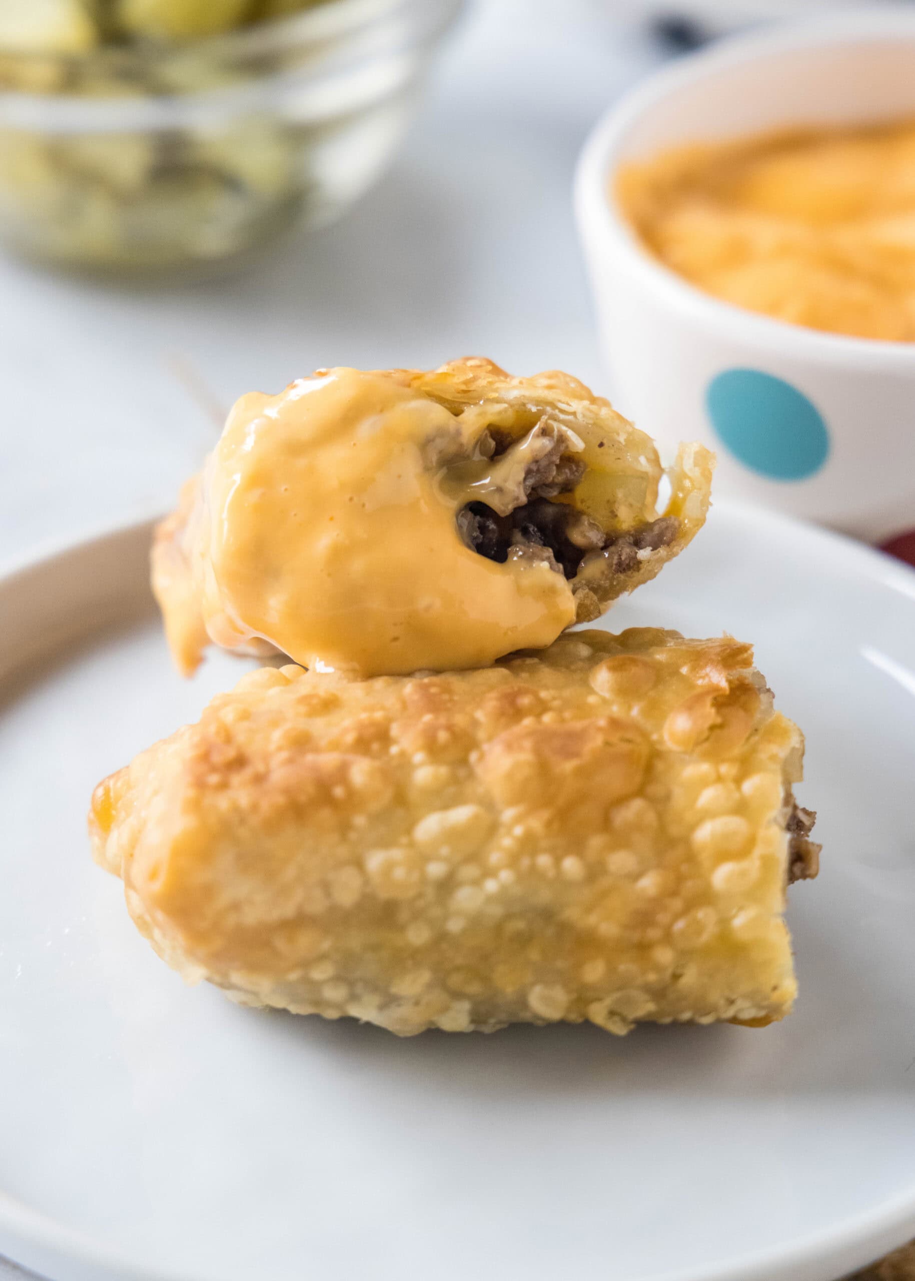 Two halves of a cheeseburger egg roll stacked on a white plate with a bowl of dipping sauce in the background.