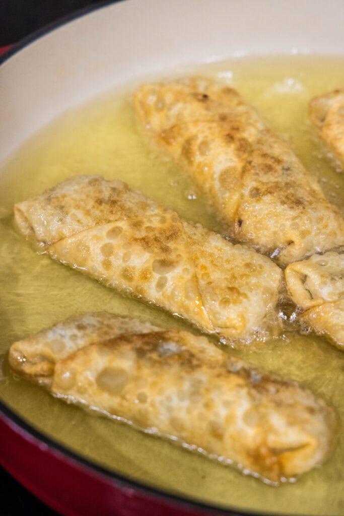 Cheeseburger egg rolls deep frying in a skillet with oil.