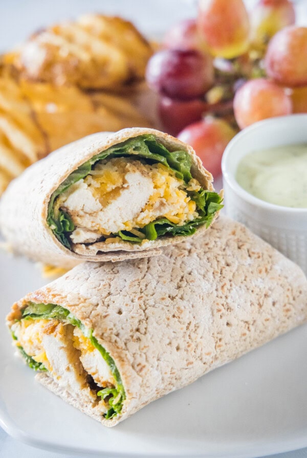 Close up of two halves of a Chick Fil A Cool Wrap on a plate next to a bowl of dipping sauce, grapes, and potato chips.