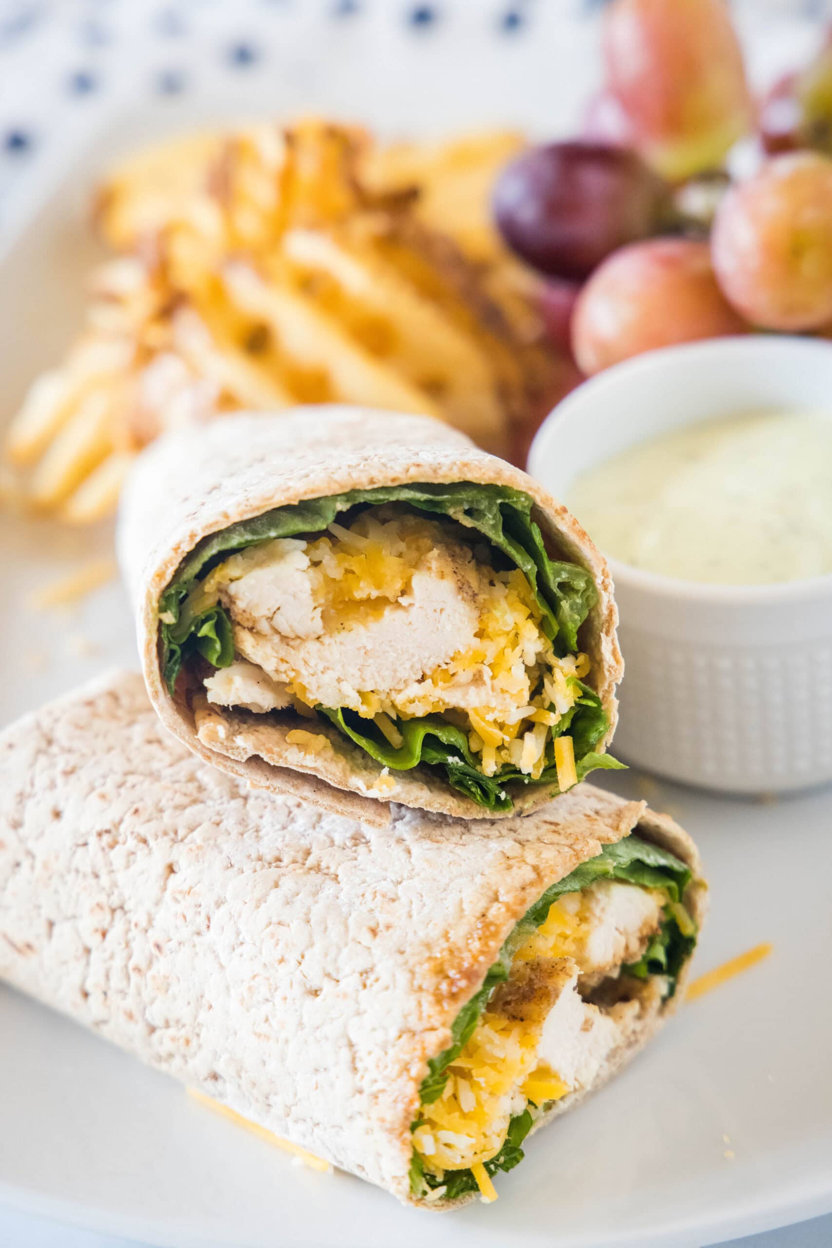 Two halves of a Chick Fil A Cool Wrap on a plate next to a bowl of dipping sauce, grapes, and potato chips.
