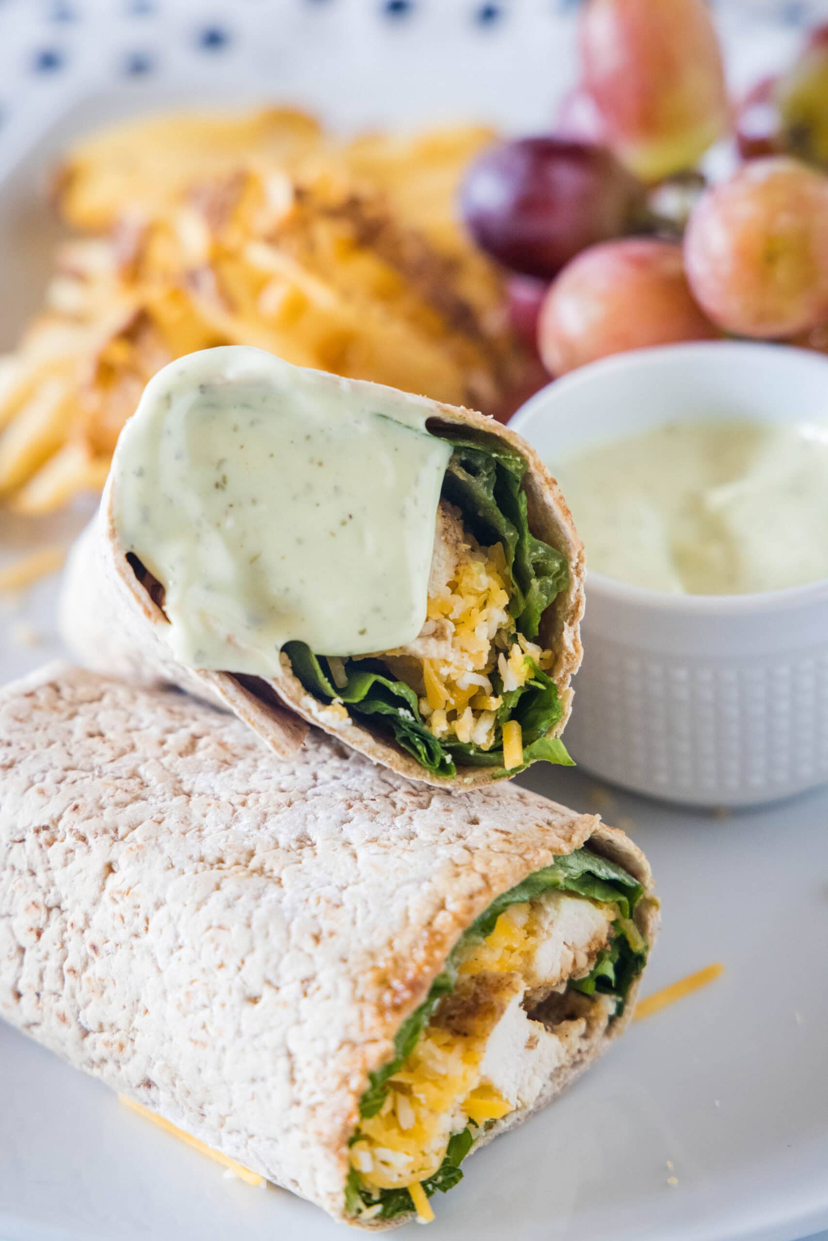 Two halves of a Chick Fil A Cool Wrap on a plate, with one half covered in dipping sauce, with a bowl of dipping sauce, grapes, and potato chips in the background.