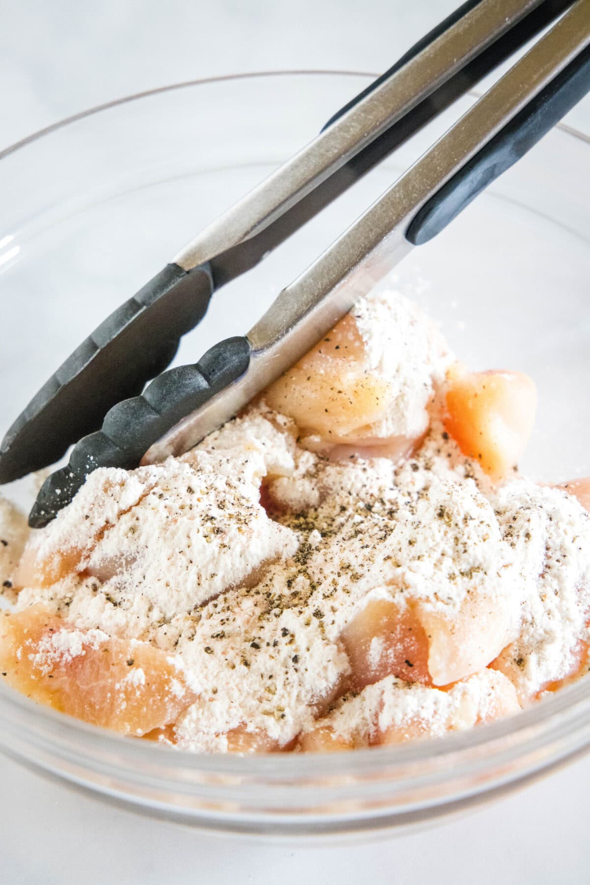 Flour added to chicken pieces in a glass bowl, with tongs.