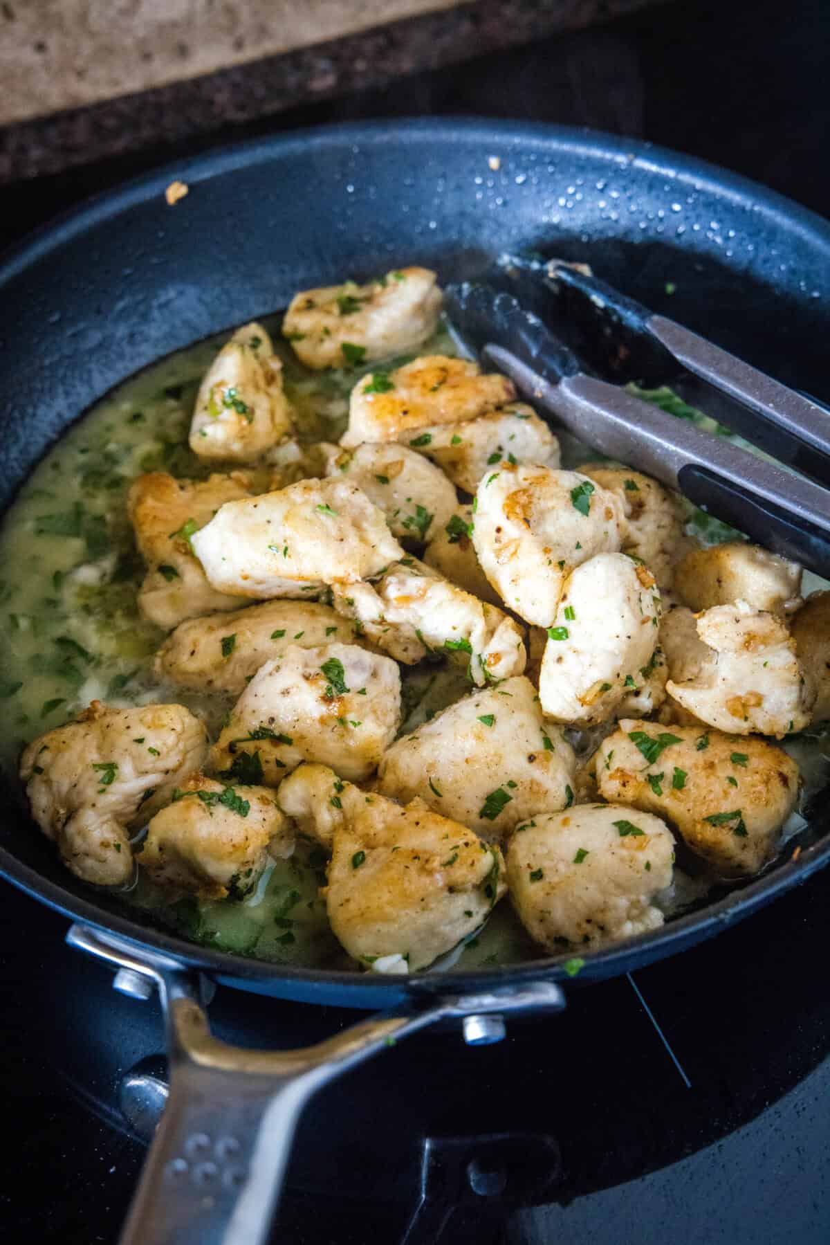 Chicken scampi in a skillet with a pair of tongs.