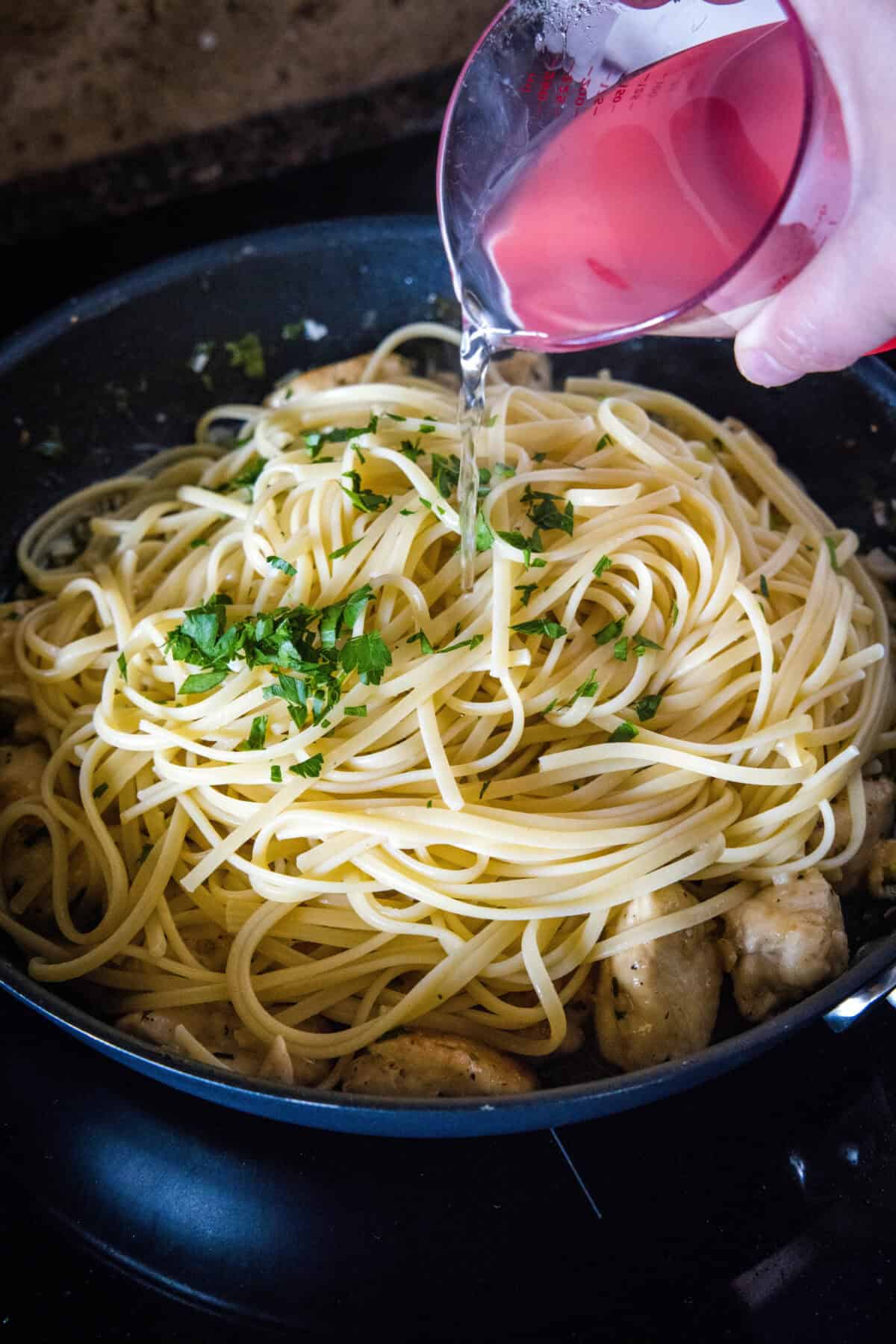 Pasta water being added to chicken scampi and cooked spaghetti in a skillet.